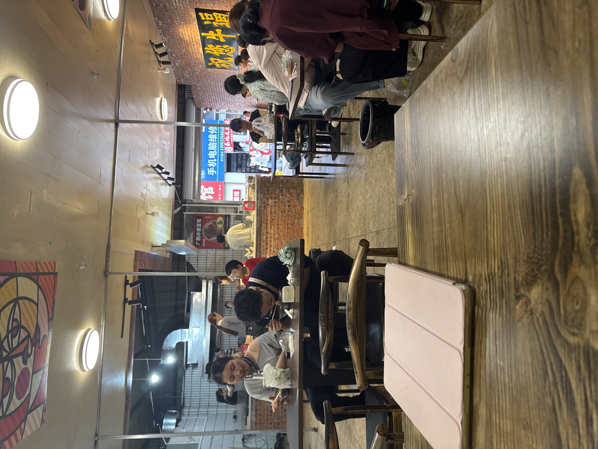 The image shows the interior of a bustling restaurant in the Nan'an District of China. The long, narrow dining area has rows of wooden tables and chairs occupied by customers eating. Overhead lighting fixtures are spaced along the ceiling.
People are seen seated at the tables, some focused on their meals, others looking at their phones. The environment includes a brick wall on one side with signage in Chinese characters, and a vibrant mural on the other wall. One sign reads, "广西冰渣豆花" (Guangxi Ice Slag Bean Curd). Another nearby advertisement says "手机电脑维修, 五公里店 13062366618" (Mobile phone computer repair, Five Kilometers shop 13062366618).
On the table closest to the camera is a wooden table top with a tablet. In the distance, activity is visible through an open area, suggesting a street scene or outdoor market. The overall impression is of a busy, local eatery with a mix of patrons and a utilitarian interior.