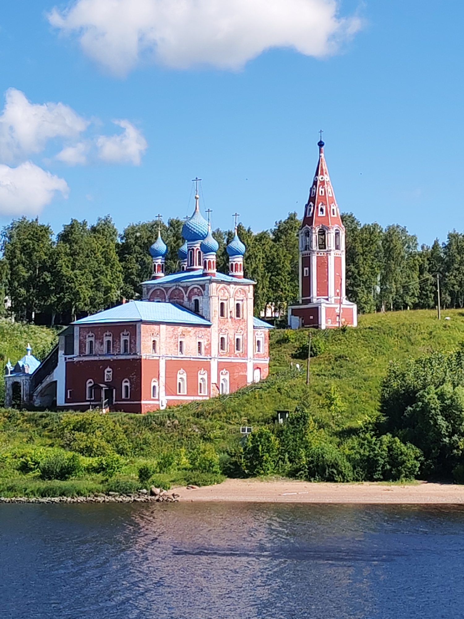 The image shows a red and white church with blue domes situated on a grassy hill overlooking a body of water. To the right of the church is a red bell tower with white accents. The church and bell tower are surrounded by a dense line of green trees. The sky is blue with scattered white clouds. The body of water in the foreground is dark blue with visible ripples. The scene appears to be a serene landscape with a prominent architectural feature. This location is the Church of the Assumption of the Blessed Virgin Mary in the city of Tutaev, Russia.