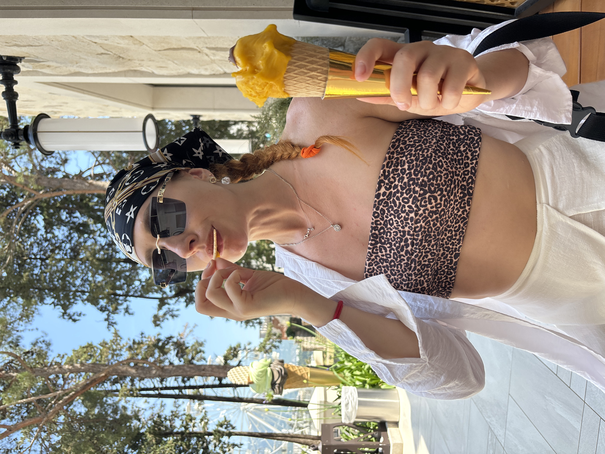 A woman stands outdoors in Budva, Montenegro, holding an ice cream cone and a small food item. She wears a black and white patterned headscarf, large sunglasses, a leopard-print bandeau top, and a white open shirt. A white skirt is visible below her waist. Her right hand holds the gold-colored ice cream cone with yellow ice cream, while her left hand brings a small food item to her mouth. The background shows trees, a clear blue sky, a building with stone cladding, a lamppost, a Ferris wheel, and a large decorative ice cream cone sculpture. The ground is paved with light-colored tiles.