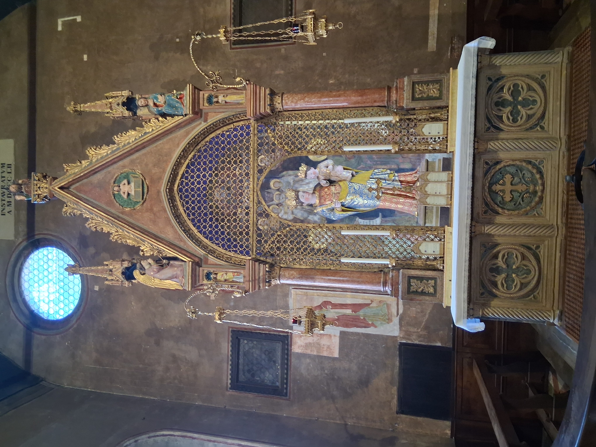 The image shows the high altar of the Basilica di Sant'Antonio da Padova in Padua, Italy. The altar is elaborately decorated with a Gothic-style tabernacle. In the center of the tabernacle is a statue of the Virgin Mary holding the Christ Child, both wearing crowns. They are surrounded by a mosaic of angels. The tabernacle is adorned with gold filigree, red marble columns, and pinnacles. Two white candles stand on either side of the statue. Above the tabernacle, a circular stained-glass window with a blue lattice pattern is visible. To the left of the altar, there is a fresco of two figures and a hanging lamp. To the right, another hanging lamp is visible. The altar itself is covered with a white cloth. The altar base is decorated with carved quatrefoil motifs. Wooden pews are visible in the foreground and to the left. The walls of the basilica are a muted brown color.