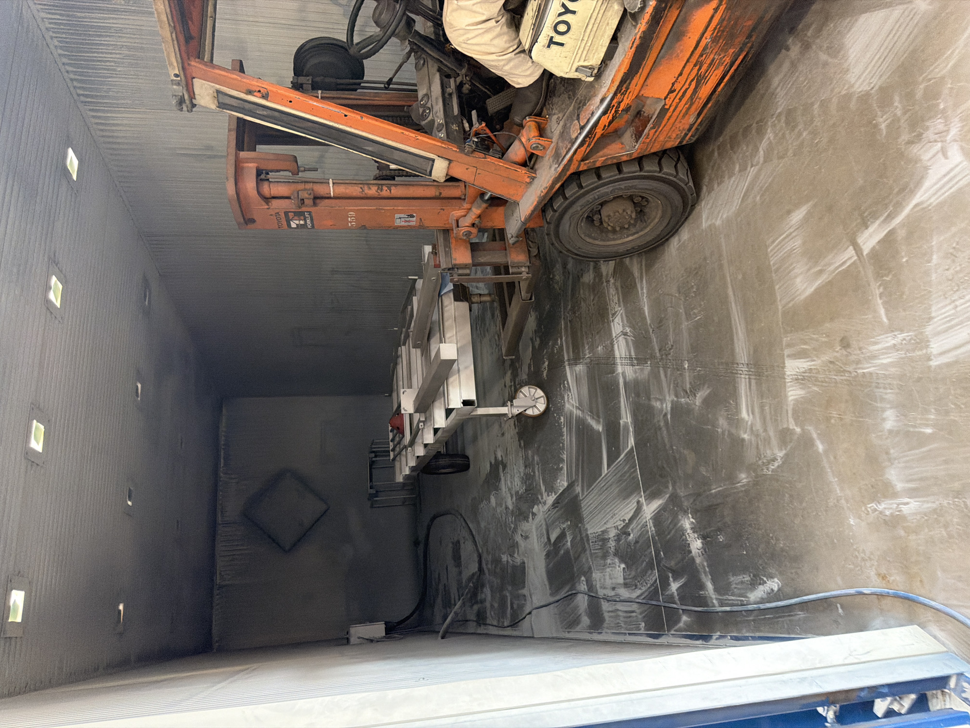 The image shows the interior of a dusty industrial workshop, likely in Mol, Belgium. An orange Toyota forklift is parked on the left side of the frame. The forklift has a person's arm resting on its console. In front of the forklift, a long, metal rack on wheels is positioned. The floor of the workshop is covered in a thick layer of white dust, with tire marks from the forklift clearly visible. The walls and ceiling are also dusty, and there are several square light fixtures on the ceiling. In the background, a diamond-shaped outline is visible on the wall. A blue hose runs along the floor, connected to a white electrical box on the left wall.
