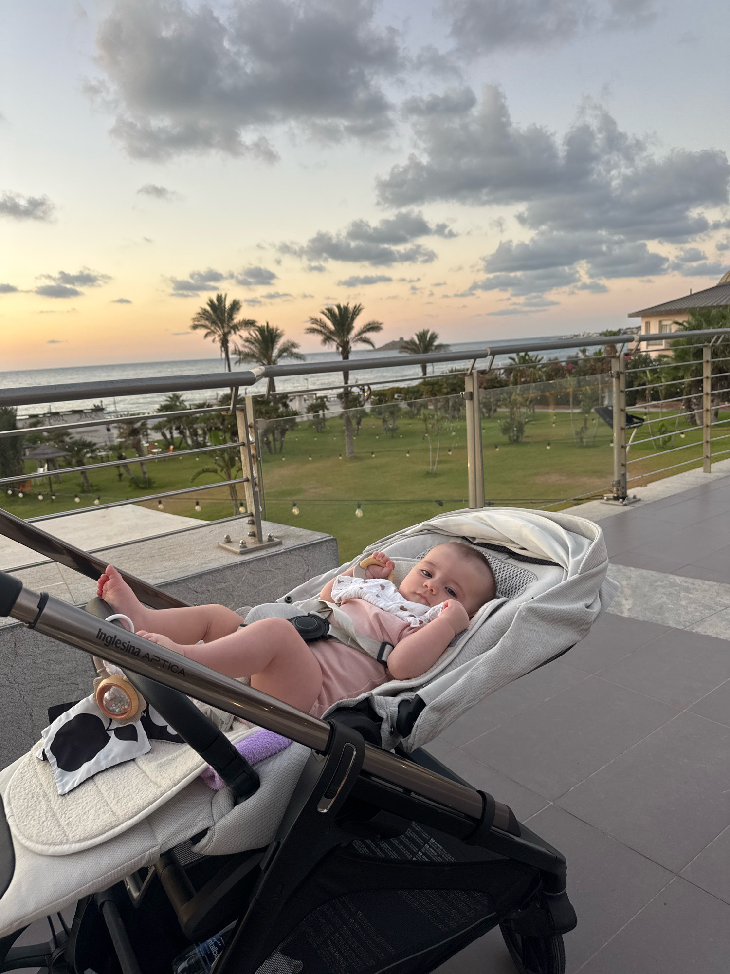 A baby is lying in a stroller on a paved patio overlooking the sea. The sky is cloudy with hints of orange and yellow from a sunset. Palm trees and a building are visible in the background. The stroller is gray and white, and the baby is wearing a pink outfit. The scene appears to be at a resort or hotel in Isola delle Femmine, Italy, given the palm trees and proximity to the sea.