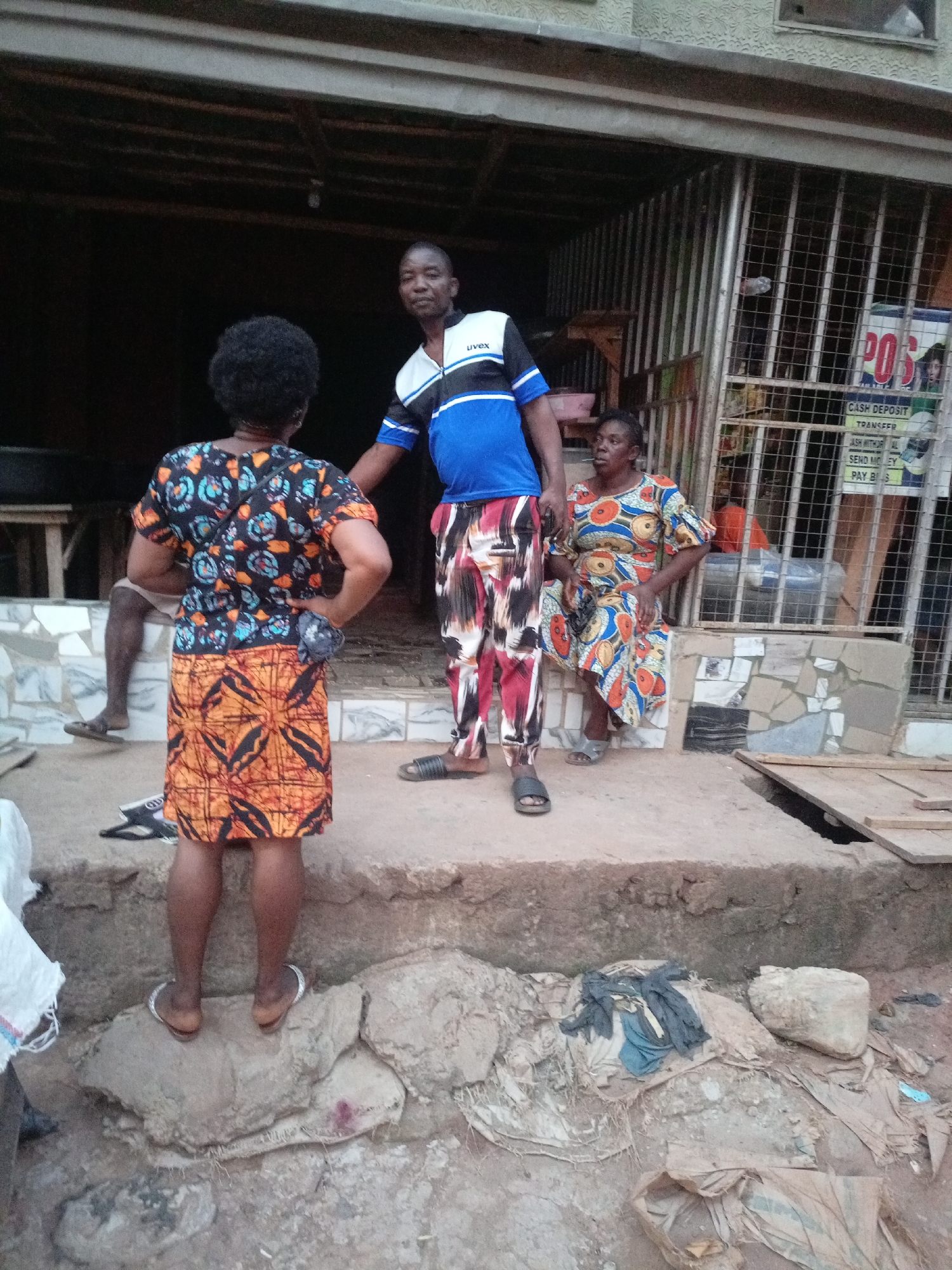 The image shows a scene in what appears to be a commercial area in Onitsha, Nigeria. Three adults are visible. A woman with short, dark hair and wearing a colorful patterned dress stands with her back to the camera. A man in a blue and white shirt and patterned pants stands facing her, with his hand near her arm. Another woman in a patterned dress sits near the entrance of a shop or stall with metal bars. A sign on the stall indicates "POS" services. The ground is uneven and appears to be a mixture of rock, dirt, and debris.