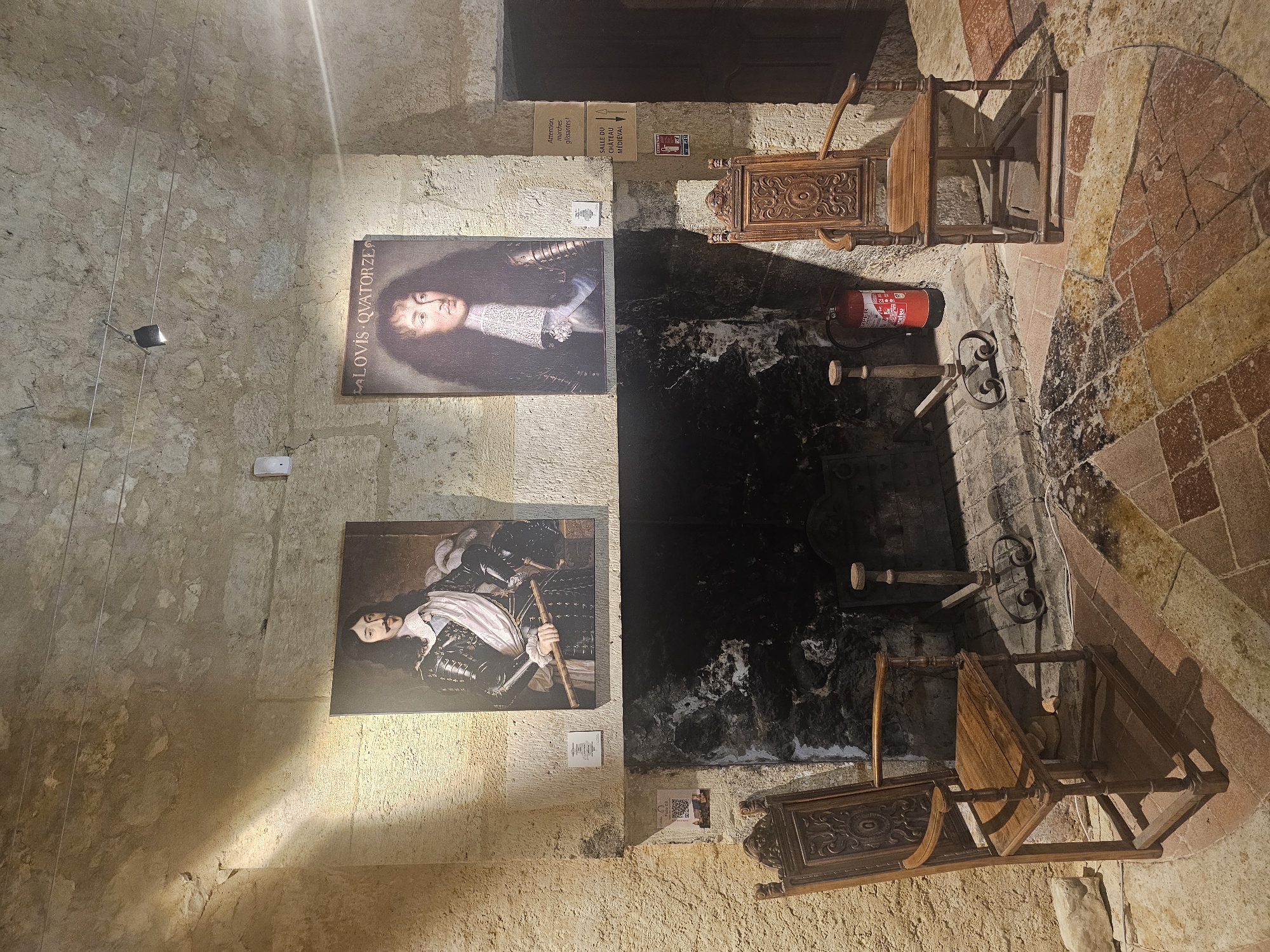 The image depicts an interior scene featuring a large stone fireplace. Two framed portraits of historical figures hang above the fireplace. The portrait on the left shows a man in armor with a sash and a baton, while the portrait on the right shows a man with long, dark hair and a ruffled collar. In front of the fireplace, two ornate wooden chairs are positioned on a patterned brick floor. A fire grate and poker are visible within the fireplace. To the right of the fireplace, a red fire extinguisher is present. A sign with text in French is affixed to the wall to the right of the right portrait, and another sign with a QR code is to the left of the left chair. The walls are made of rough-hewn stone, and a spotlight illuminates the scene from above. The scene appears to be in a historical building or museum.