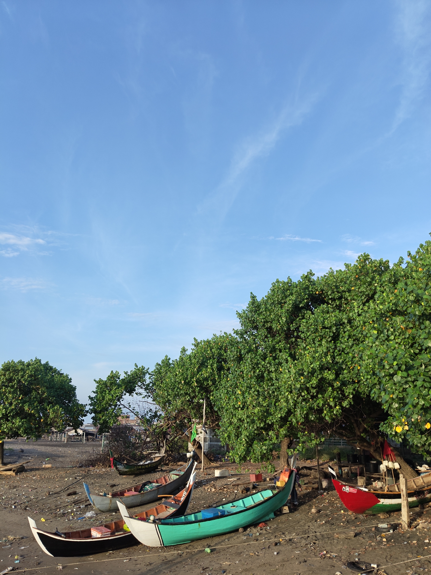 The image captures a coastal scene in Lhokseumawe, Indonesia. Several small fishing boats are beached on dark sand. The boats are painted in various colors, including black, white, green, and red. Behind the boats are trees with dense green foliage. Debris and litter are scattered on the sand. In the background, there are glimpses of buildings and structures. The sky is blue with a few scattered clouds.