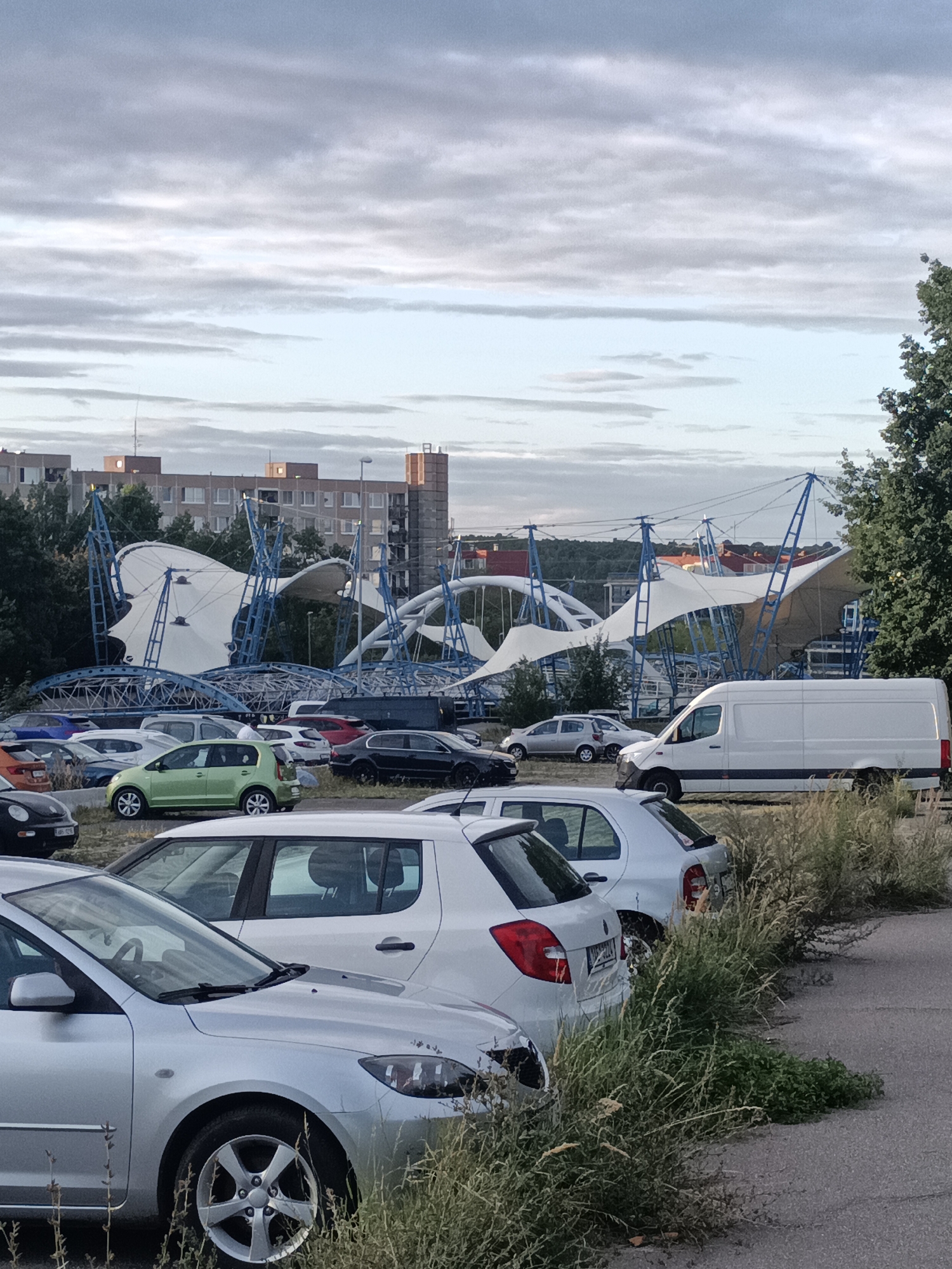 A parking lot is filled with various vehicles. Among the cars, there is a white van. In the background, a building with a unique architectural design featuring a large white tent-like structure supported by blue metal poles is visible. The sky is cloudy. The scene appears to be an outdoor setting in a city.