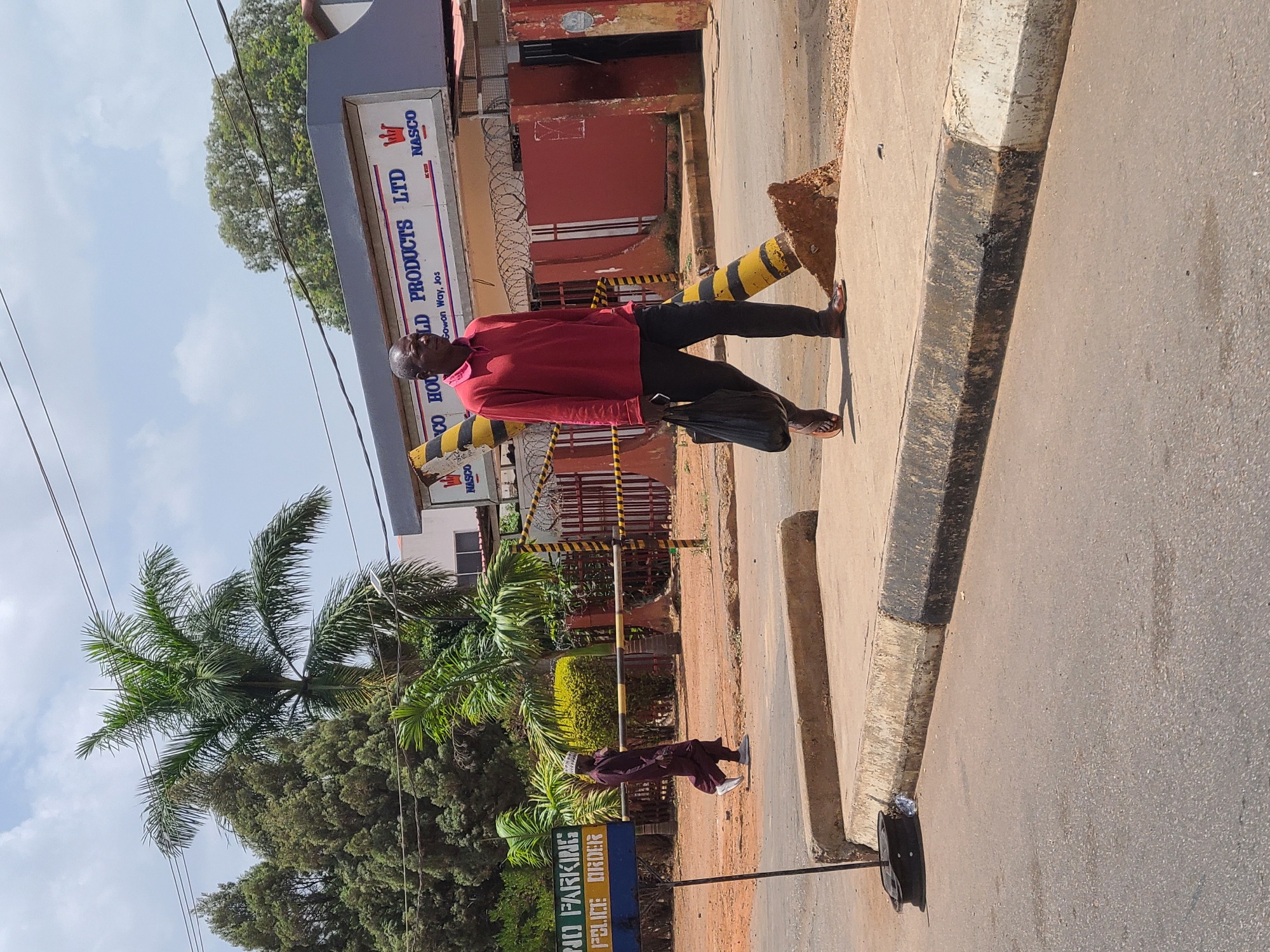 The image is taken on Gowon Way, Jos, Nigeria. A man wearing a red long-sleeved shirt and dark pants is walking along the sidewalk. He is carrying a black bag in his right hand and is wearing sandals. Behind him is a building with a sign that reads "NASCO HOUSEHOLD PRODUCTS LTD." There is a security fence with barbed wire surrounding the building. Yellow and black striped bollards are in front of the building. On the other side of the sidewalk are trees and a sign that reads "NO PARKING POLICE ORDER". Another man is standing behind the trees on the sidewalk. The sky is partly cloudy.