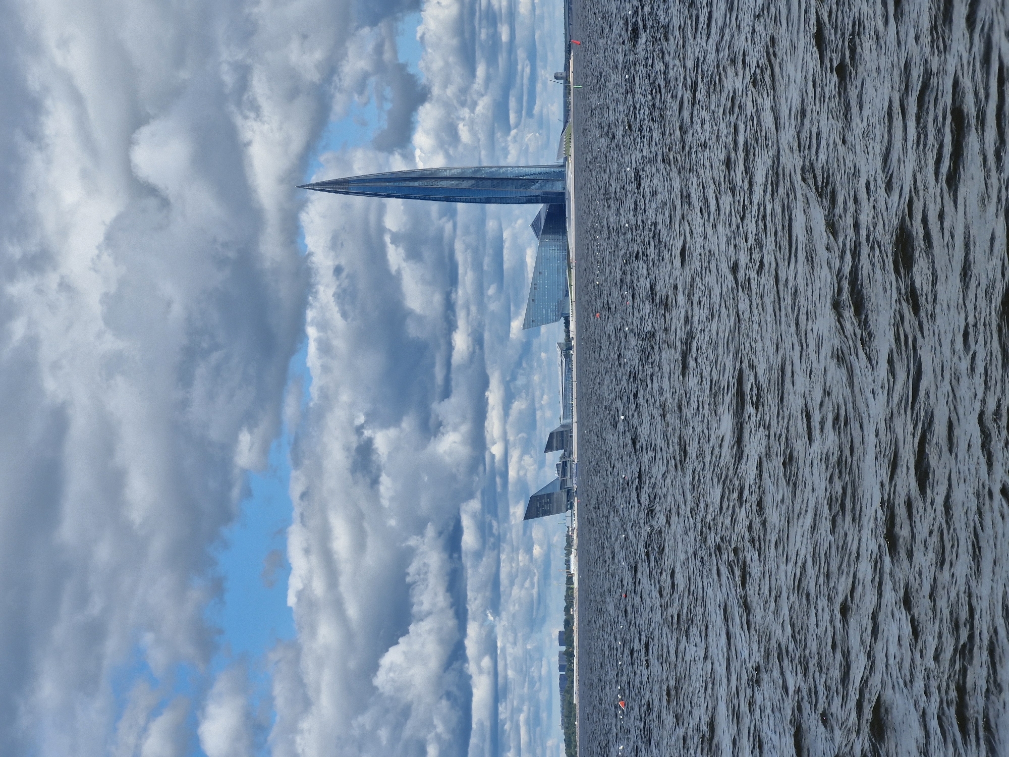 A wide shot captures the Lakhta Center skyscraper in Saint Petersburg, Russia, as a dominant focal point, its sleek, twisting glass exterior reflecting the cloudy sky. The skyscraper is situated on the Neva Bay, and the foreground is filled with dark, choppy water, dotted with numerous small, white buoys and a few orange markers. In the middle ground, a modern complex of buildings with sharp angles and glass facades, including the Lakhta Center, is visible on the shoreline. Further in the distance, a treeline and urban structures are discernible. The sky is a dramatic canvas of heavy, gray clouds with patches of bright blue sky peeking through, suggesting a dynamic weather pattern.