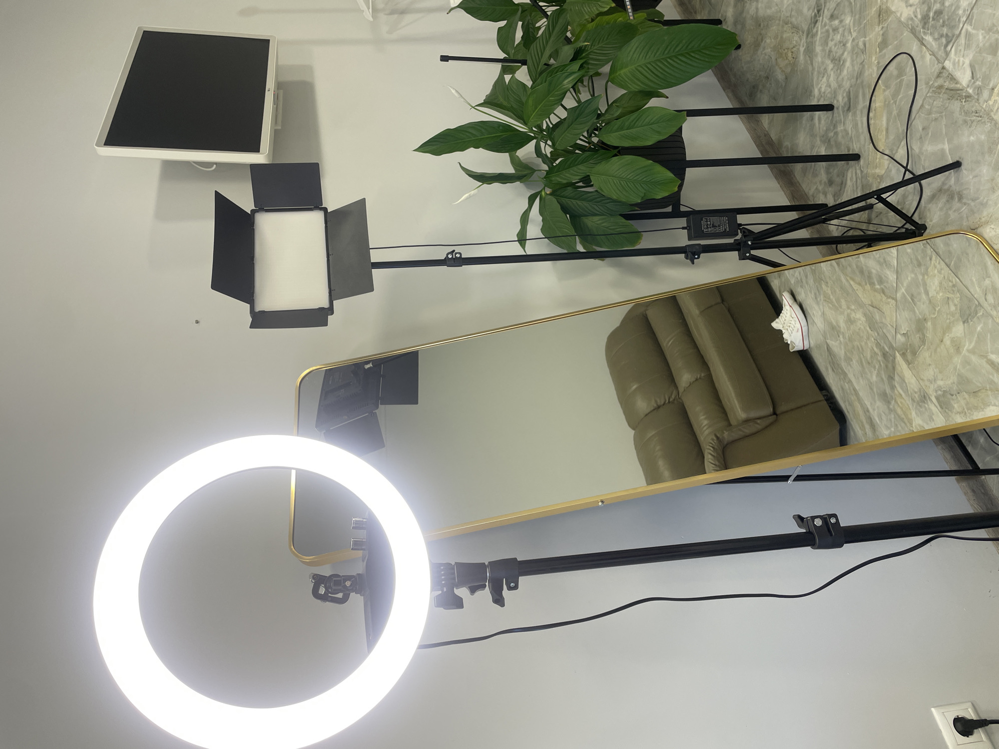 The image displays an indoor setting, likely a dedicated space for photography or video recording, featuring various pieces of equipment. The room has light grey walls and a grey marble-patterned floor.
In the foreground, a large, circular LED ring light, brightly illuminated, is mounted on a black stand. To its right, another professional-grade rectangular LED panel light with adjustable black barn doors is positioned on a separate stand. Further back and slightly to the left, a flat white monitor or light panel is affixed to the wall.
A tall, gold-framed full-length mirror stands vertically, reflecting a comfortable-looking olive-green or tan leather recliner sofa or chair, and a section of the marble floor. A pair of white low-top sneakers, possibly Converse, with white laces and a subtle red stripe on the sole, is resting on the floor near the mirror, partially visible directly and in reflection.
Behind the rectangular light, a large potted peace lily plant with vibrant green leaves and several white blossoms is situated on a black metal stand, adding a natural element to the setup. Black electrical cables are visible running across the floor and along the light stands. A white European-style power outlet with a black plug inserted is on the lower right wall.
Small, unreadable white text is present on a black power adapter for one of the lighting units. The arrangement of equipment suggests an environment prepared for content creation, such as vlogging, live streaming, or product photography. The setting appears clean, modern, and well-lit by artificial sources.