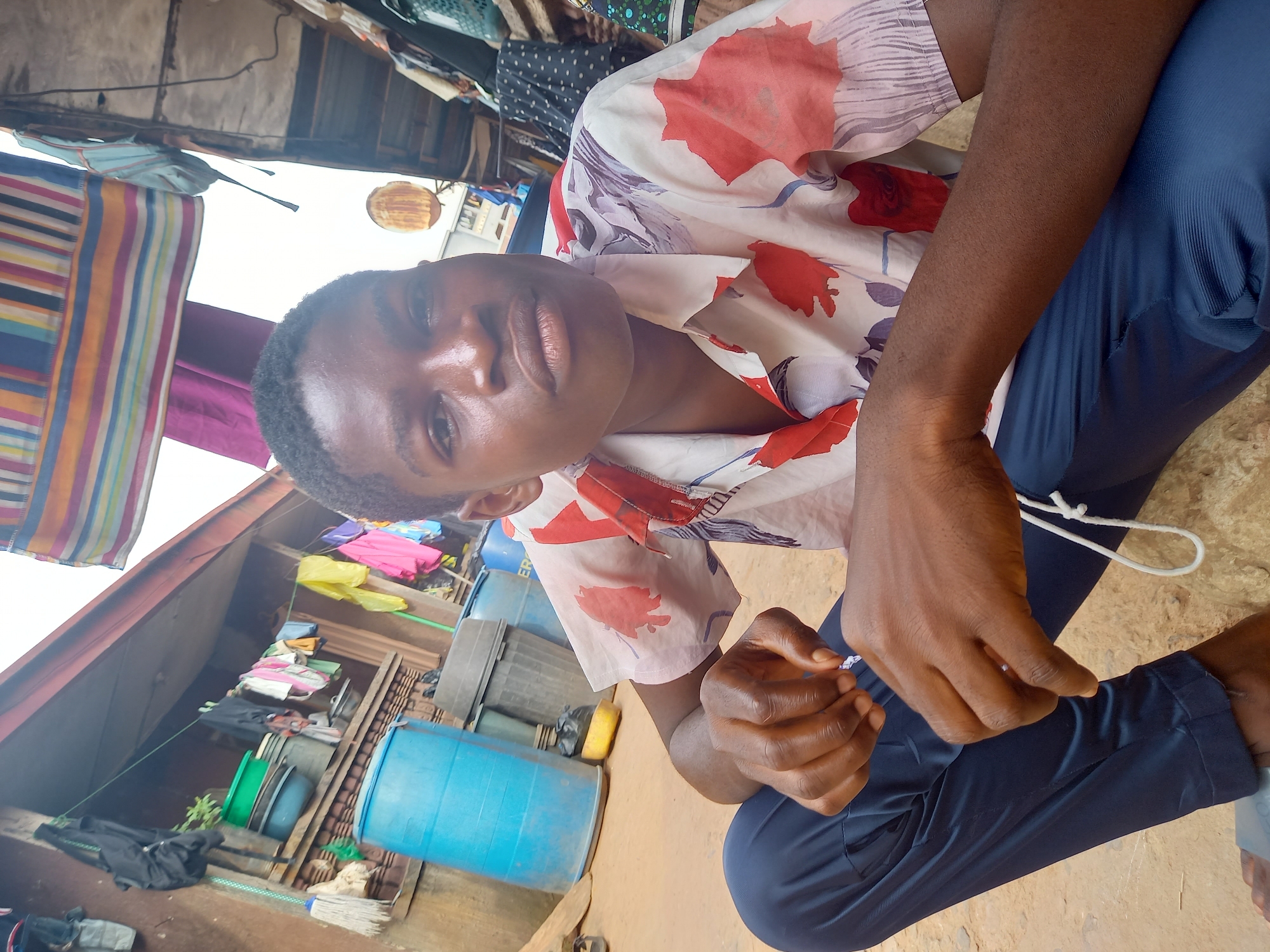 A young individual, appearing male, is seated on the ground and looks directly at the camera with a neutral expression. He has dark skin and is wearing a light-colored, short-sleeved shirt adorned with red, pink, and purple floral patterns, paired with dark blue trousers. His hands are clasped in front of him, holding or manipulating a thin white string or cord.
The setting is an outdoor, somewhat informal environment, likely a yard or a commercial area in Onitsha, Nigeria. In the background, several large plastic barrels are visible, including a bright blue one and a dark gray one, suggesting water storage or utility use. Various household items, such as pots, basins, bags, and clothes, are scattered or stacked around, alongside a broom. The surrounding structures appear to be part of a building or a makeshift shelter with corrugated metal or wooden elements.
Above the scene, a bright white sky indicates it is daytime. Clothes are hanging on a line, including a colorful striped fabric and a purple cloth. A rusty, round metal object is visible hanging in the upper background. The overall impression is one of a practical, everyday local setting. No visible text is clearly discernible in the image.