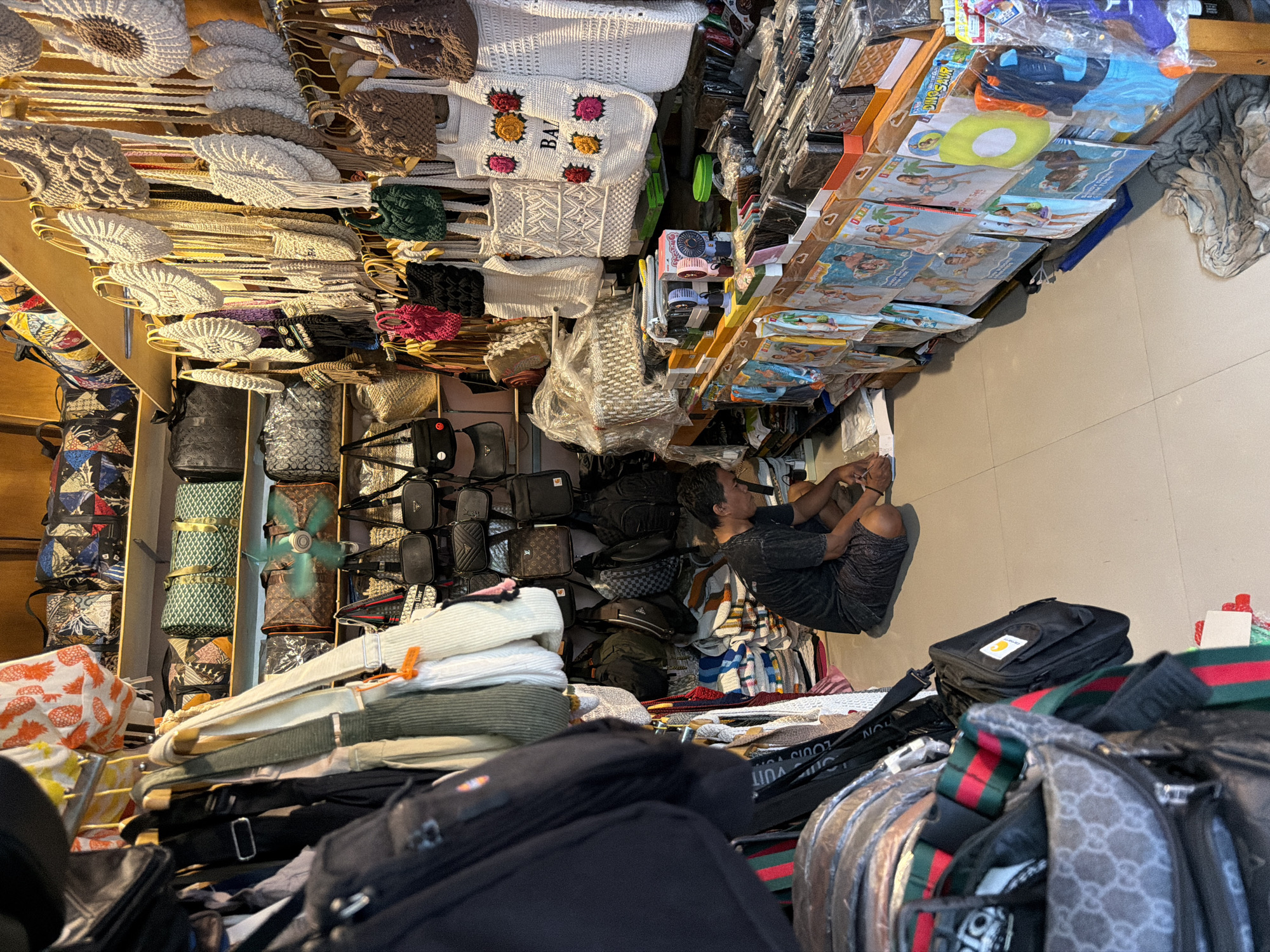 A man is sitting on the floor in what appears to be a small retail shop in North Kuta, Indonesia. The shop is filled with merchandise, predominantly bags and accessories, displayed on shelves and hanging from racks. The man is dressed in a dark t-shirt and shorts and is looking down at some papers on the floor in front of him, appearing to be working. The shop is packed with various types of bags, including tote bags, handbags, backpacks, and clutches, made from different materials like woven straw, crochet, and leather. Some bags are decorated with colorful patterns or designs, while others are plain. In the background, there are shelves stocked with more bags and other items. A small electric fan is visible on a shelf, possibly for cooling the shop. The floor is tiled and clean. The overall impression is of a busy, well-stocked shop selling a variety of fashion accessories.