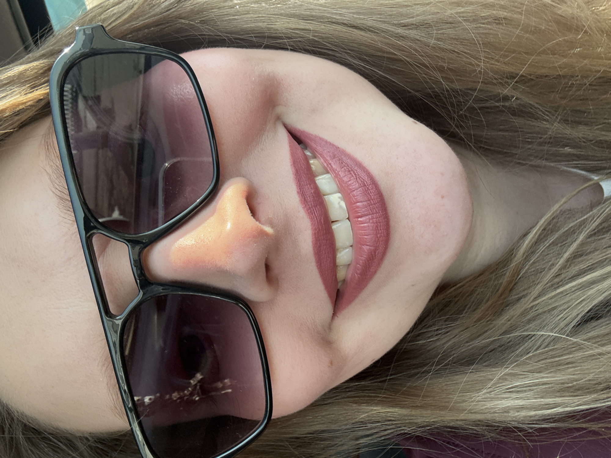 A close-up of a smiling woman's face. She is wearing dark sunglasses with a rectangular frame. Her hair is blonde. She is wearing a mauve-colored lipstick. No location can be determined based on the visual information.