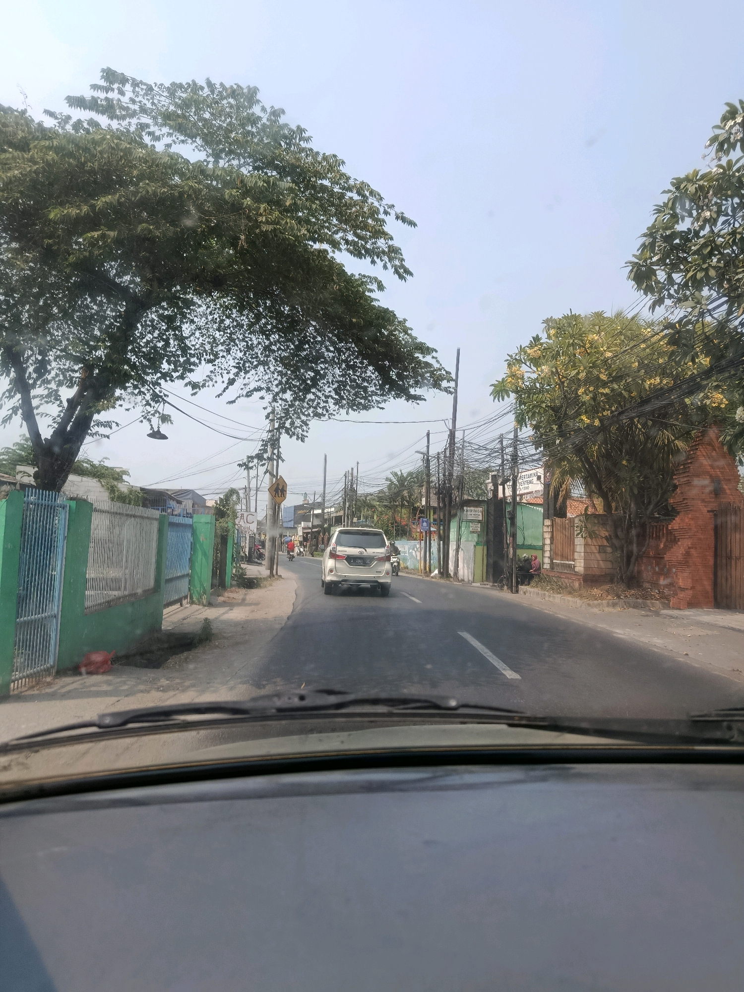 The image shows a street scene in the city of East Jakarta, Indonesia, likely during the daytime. The perspective is from inside a vehicle, looking out through the windshield. A white SUV is driving away from the camera down the asphalt road, which has a white line marking the lane. On the left side of the road, there is a green and blue fence with metal bars, and a large tree with lush green foliage overhangs the fence and the street. Further down the road on the left, there are some buildings and more fences. On the right side of the road, there is a brick wall with a decorative opening, and behind it, a tree with yellow flowers. There are numerous utility poles and wires lining the street. In the distance, there are more buildings and some pedestrians. The sky is a clear light blue, indicating a sunny day. The overall impression is of a typical urban street in a tropical climate.
