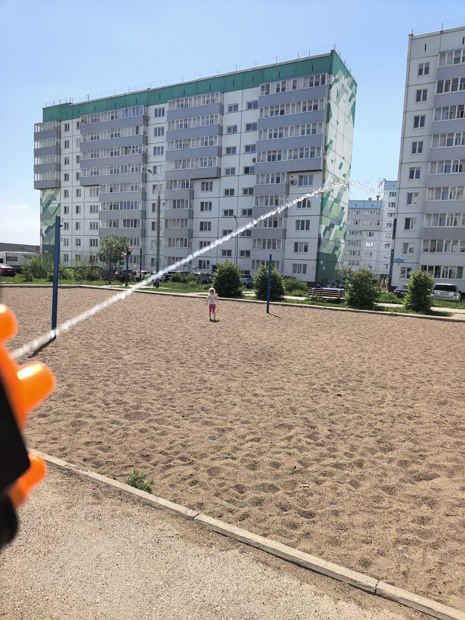 The image depicts a sand-covered playground in Фролы, Russia, in front of apartment buildings. A young girl stands in the middle of the playground. Water arcs across the playground between two blue poles. The nozzle of a water gun is visible in the lower left of the frame. The day is sunny, with a clear blue sky.