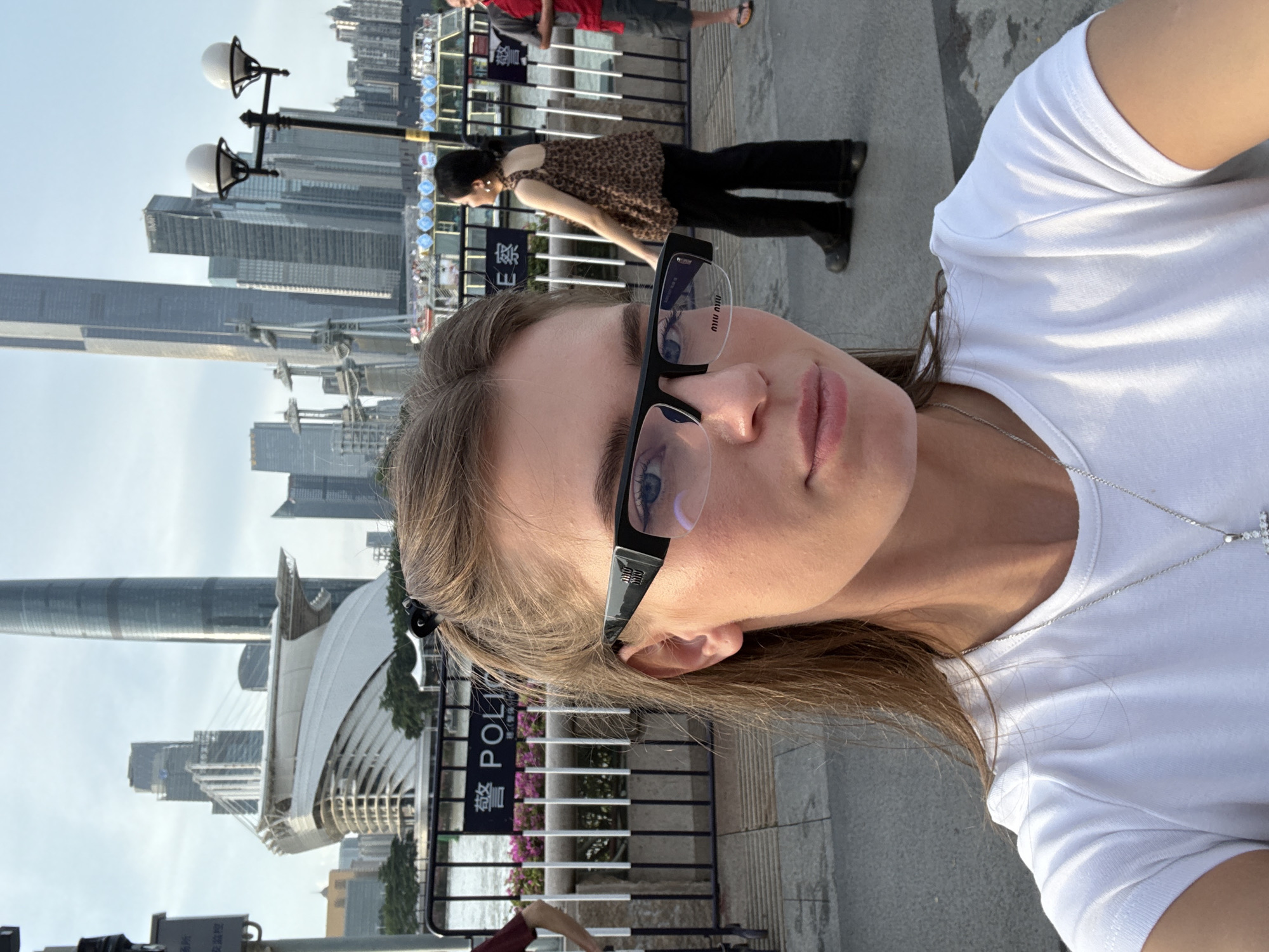 A young woman, wearing a white t-shirt, glasses, and a silver necklace with a cross pendant, takes a selfie. She is looking towards the camera, her face partially obscured by her glasses. The background shows a cityscape with modern buildings, some tall skyscrapers, and a bridge. There's a river or body of water in the foreground with some greenery and flowering plants. A black metal fence with white posts is visible, behind which a sign that appears to say "POLICE" in English, along with Chinese characters, can be seen. Other people are visible in the background, including a woman in a leopard print top and black pants, and another person in a red shirt. The lighting suggests it is daytime, likely sunny. The scene is outdoors, possibly a promenade or park area along a waterfront in an urban setting. The overall mood is casual and relaxed.