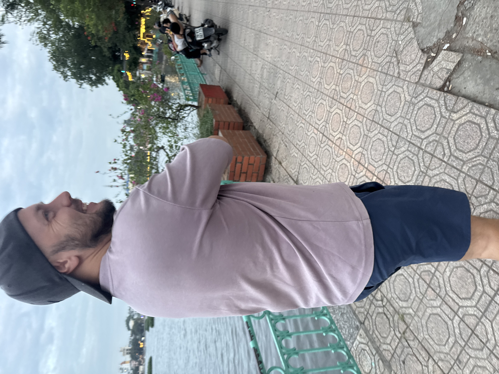 A man wearing a grey baseball cap, a mauve t-shirt, and dark blue shorts stands on a patterned tiled sidewalk next to a body of water. He is facing away from the camera, looking over his shoulder and smiling. The water is bordered by a green metal railing, and behind it, several trees with pink flowers are visible. Further in the background, buildings and lights suggest an urban setting. Several motorcycles are parked along the sidewalk, and two people are resting on one of them. The sky is overcast, indicating it is likely early morning or late evening. The overall scene depicts a relaxed atmosphere in a park or along a waterfront area in Hanoi, Vietnam.