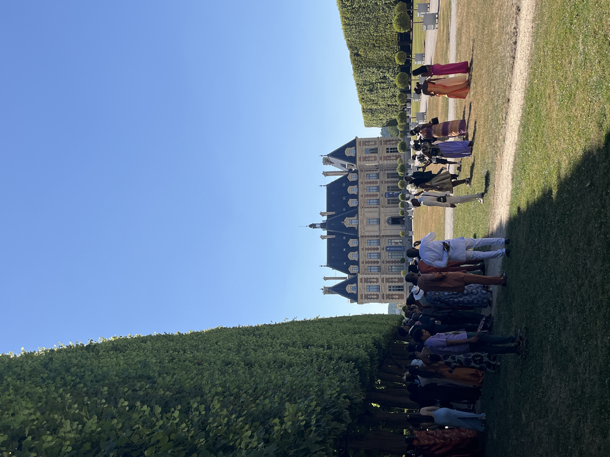 This is a vertical, eye-level, full-shot photograph taken outdoors on a sunny day. The scene depicts a gathering of people in front of a large, classical French chateau, likely in the city of Sceaux, France, given its resemblance to the Chateau de Sceaux. The chateau is a stately building with a mansard roof, multiple chimneys, and numerous windows. It is framed by two very tall, manicured green hedges that extend from the foreground towards the chateau. The hedge on the left is much closer to the viewer and runs along the left edge of the frame, creating a strong vertical line. The hedge on the right is further away, creating a softer, less defined boundary on that side.
A diverse group of people, dressed in what appears to be celebratory attire, are gathered in the middle ground and foreground. Many are wearing colorful garments, including dresses and suits. Some are standing in small groups, while others are more spread out. Their postures suggest they are socializing or posing for photographs. The ground is covered in a mixture of green grass and dry, yellowed grass, indicating a well-maintained but possibly dry landscape. In the background, beyond the chateau, the sky is a clear, vibrant blue. Small, round trees are visible in neat rows in front of the right hedge.