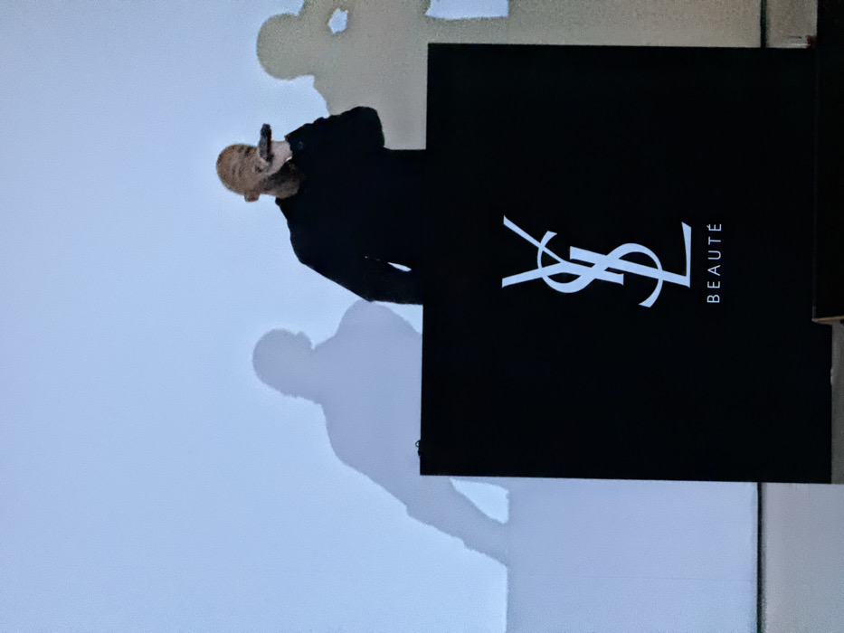 A man in black holds a microphone and stands beside a black display with a white YSL logo and the word "BEAUTÉ" vertically arranged on the right. The scene is cast in a cool light with shadows of the man and a blurred figure on the background wall. The image is vertically oriented.