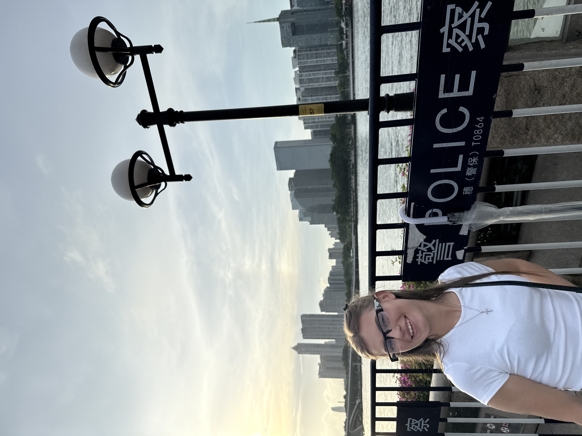 An outdoor, medium-shot, eye-level, full-body photograph features a young Caucasian woman with blonde hair and glasses smiling at the camera. She is wearing a white t-shirt and a silver cross necklace, and a black strap is visible over her right shoulder. She is standing behind a black metal fence. In the background, a cityscape with numerous modern skyscrapers is visible across a body of water. A lamppost with two spherical lights stands to the right of the woman. A dark blue sign with white lettering reads "POLICE" in English, with Chinese characters above and below the word. The location is identified as Haizhu District, China. The sky is overcast with diffused sunlight.