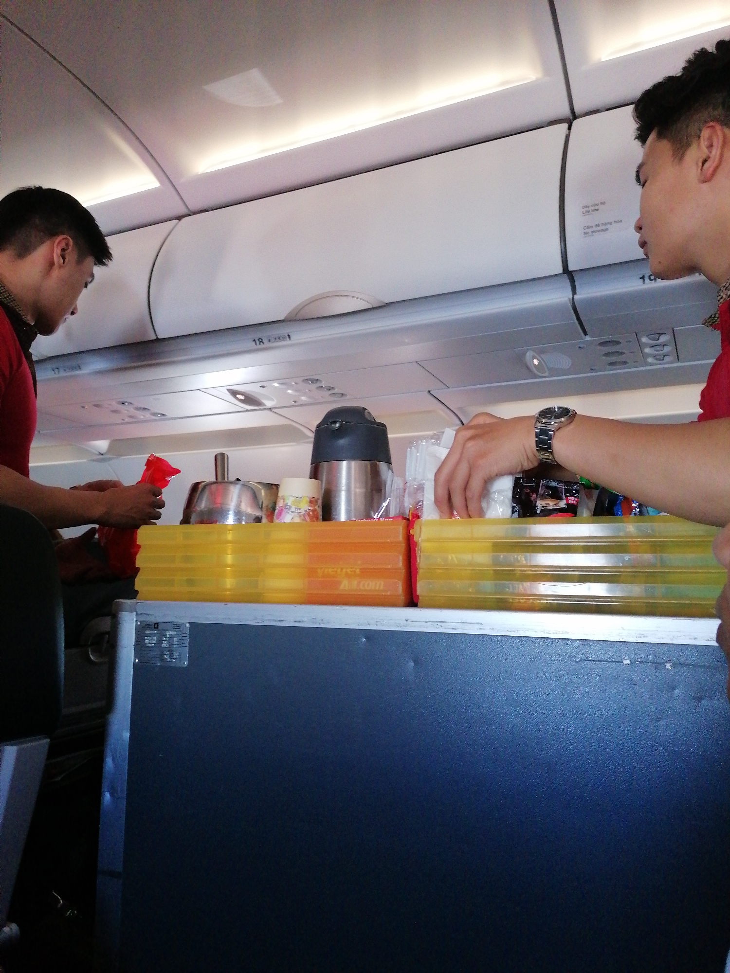 Two flight attendants in red shirts are preparing a snack and drink cart in the aisle of an airplane. The cart is dark blue with two yellow trays on top containing various items. Above the attendants, luggage compartments are visible with row numbers labeled. The location is an airplane cabin of an unknown airline, flying over an unknown city.