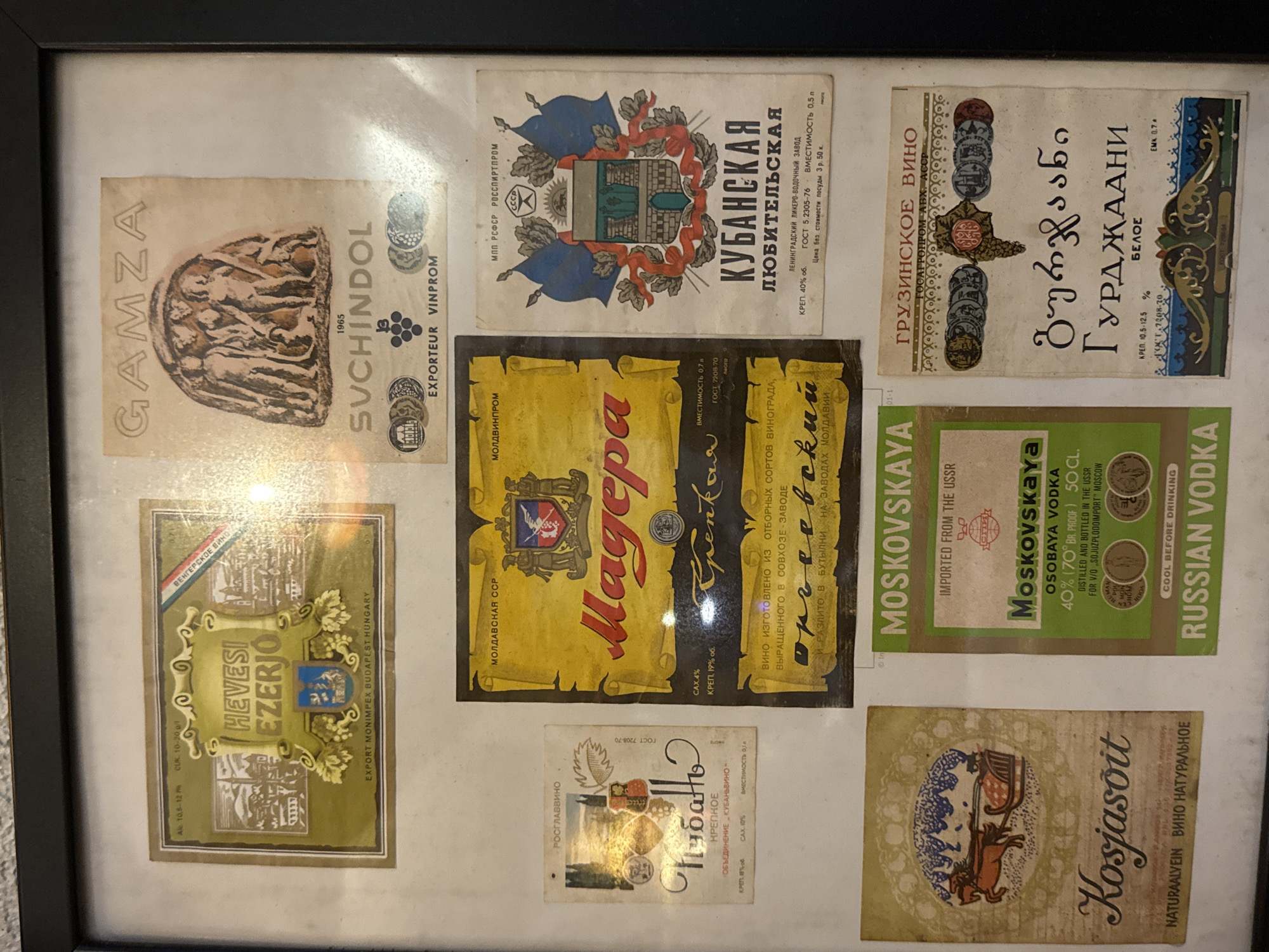 The image shows a framed collection of vintage liquor labels. The labels feature brands like "Gamza", "Svchindol", "Kubanskaya", "Hevesi Ezerjo", "Magera", "Moskovskaya", "Kosjasoit", and "Gurdzhani". The text on the labels is in various languages, including Russian, Hungarian, and Georgian. The labels are arranged on a white background within a black frame. There is no indication in the image of the city of Baan Chaweng Noi, Thailand.