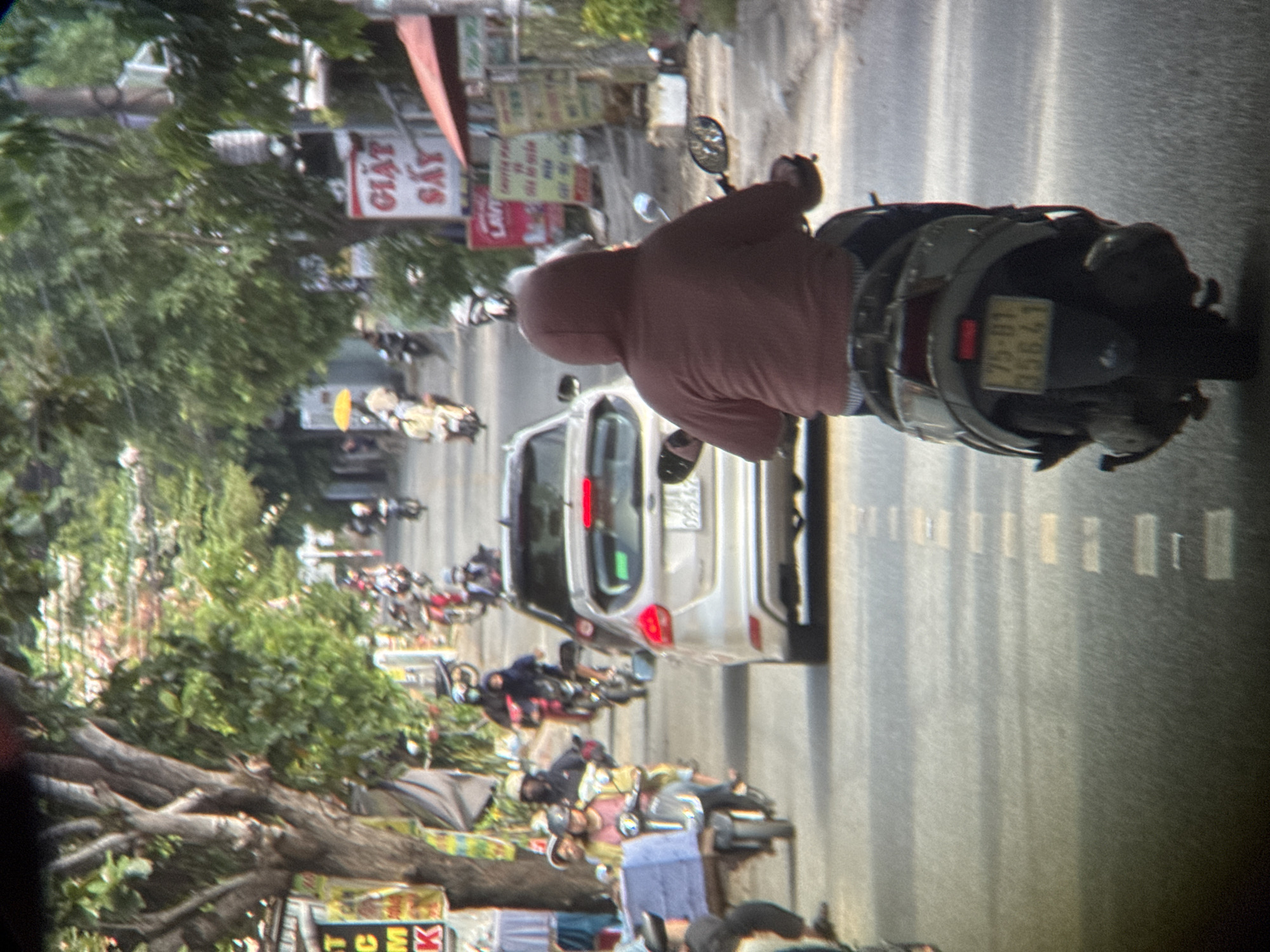 This is a medium-angle, eye-level shot of a busy street in An Cựu Ward, Vietnam. The scene is dominated by a line of motorcycles and a silver car navigating a paved road lined with trees and buildings. In the foreground, a person wearing a maroon hooded sweatshirt rides a scooter, their back to the camera. The scooter has a yellow license plate with the numbers "75-01 356.41". The car in front of the scooter is silver and appears to be a hatchback. Further down the street, other motorcycles are visible, some with riders wearing helmets. The environment suggests a sunny day, with shadows cast on the road. Buildings with signs in Vietnamese are visible on the right side of the street, including one with red lettering that reads "GIẶT SẤY". Trees with lush green foliage line both sides of the road. The overall impression is a snapshot of daily life and traffic on a Vietnamese street.