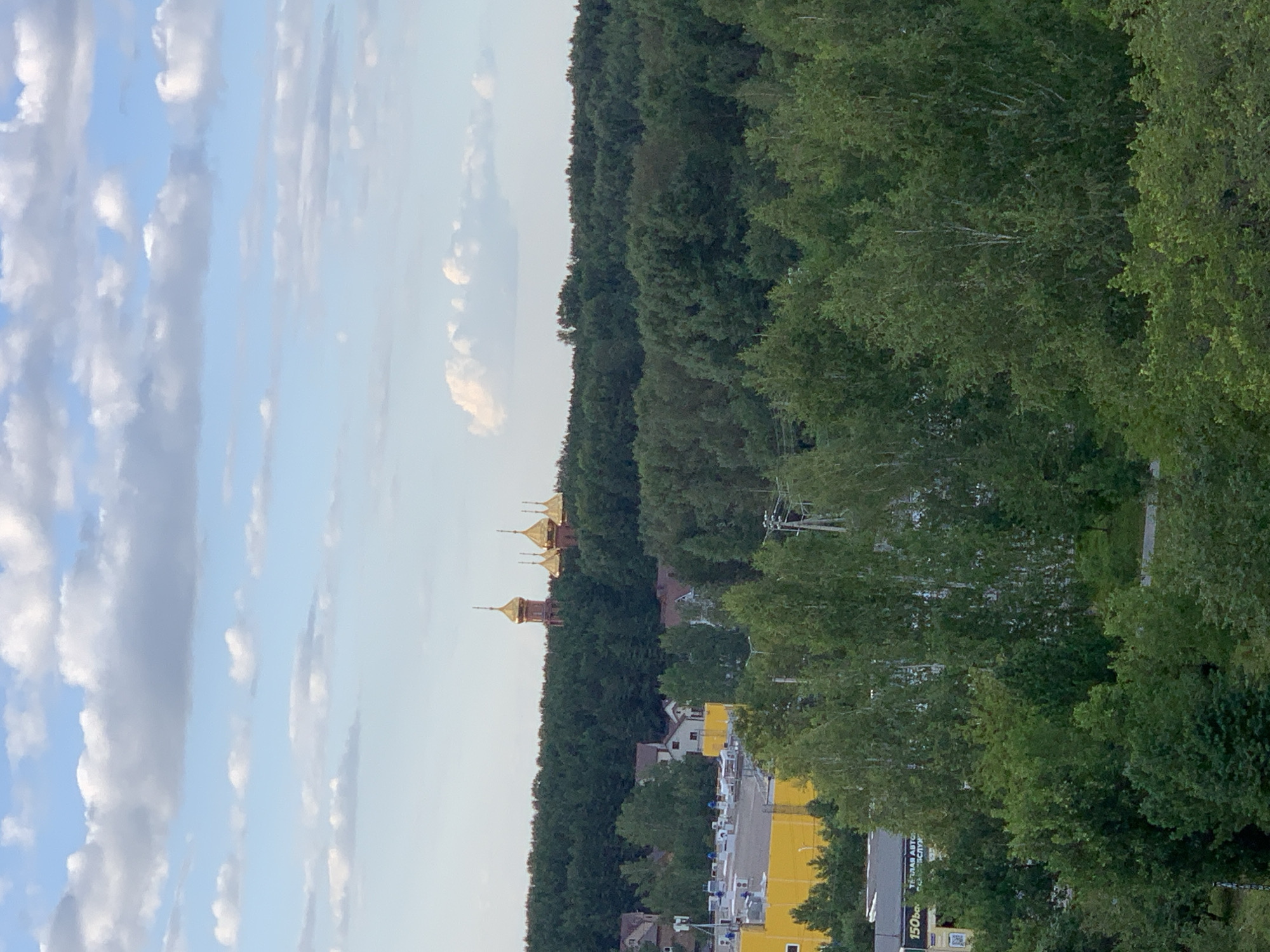 The image captures an aerial view of a wooded area with several structures visible. Dominating the background are multiple golden-domed religious buildings, possibly a monastery or church, nestled on a hill amidst a dense forest. In the mid-ground, a large yellow building with a flat roof, likely a commercial or industrial facility, is partially obscured by trees. Adjacent to it are residential houses. The foreground consists of a thick canopy of green trees. The sky above is a pale blue with scattered white clouds. No people are visible in the frame. The scene suggests a semi-urban or suburban environment where nature and man-made structures coexist.