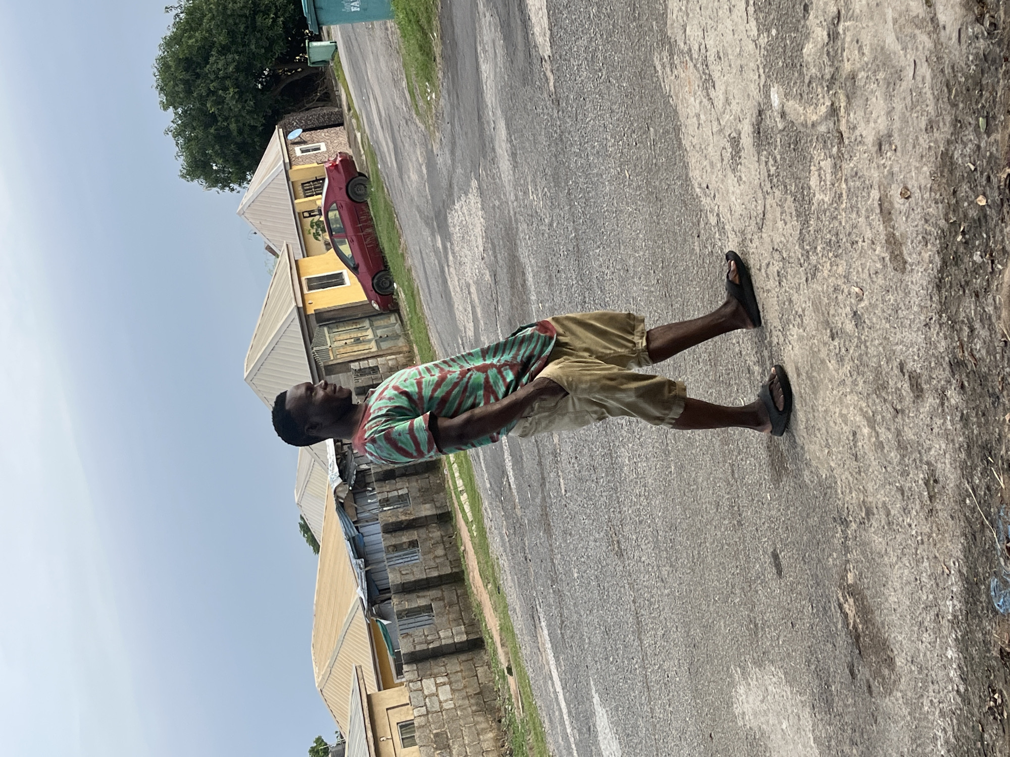 A full-body shot captures a Black man standing on a paved street in Abuja, Nigeria, in what appears to be a residential area. The man, wearing a green and red striped t-shirt and tan shorts, has his hands in his pockets and is looking to his right, away from the camera. He is wearing black flip-flops. Behind him, several small, single-story homes with light-colored roofs are visible, along with a red sedan parked on the side of the road. The sky above is overcast. A green trash bin is also visible to the right of the frame. The overall scene is that of an everyday street in a Nigerian city.