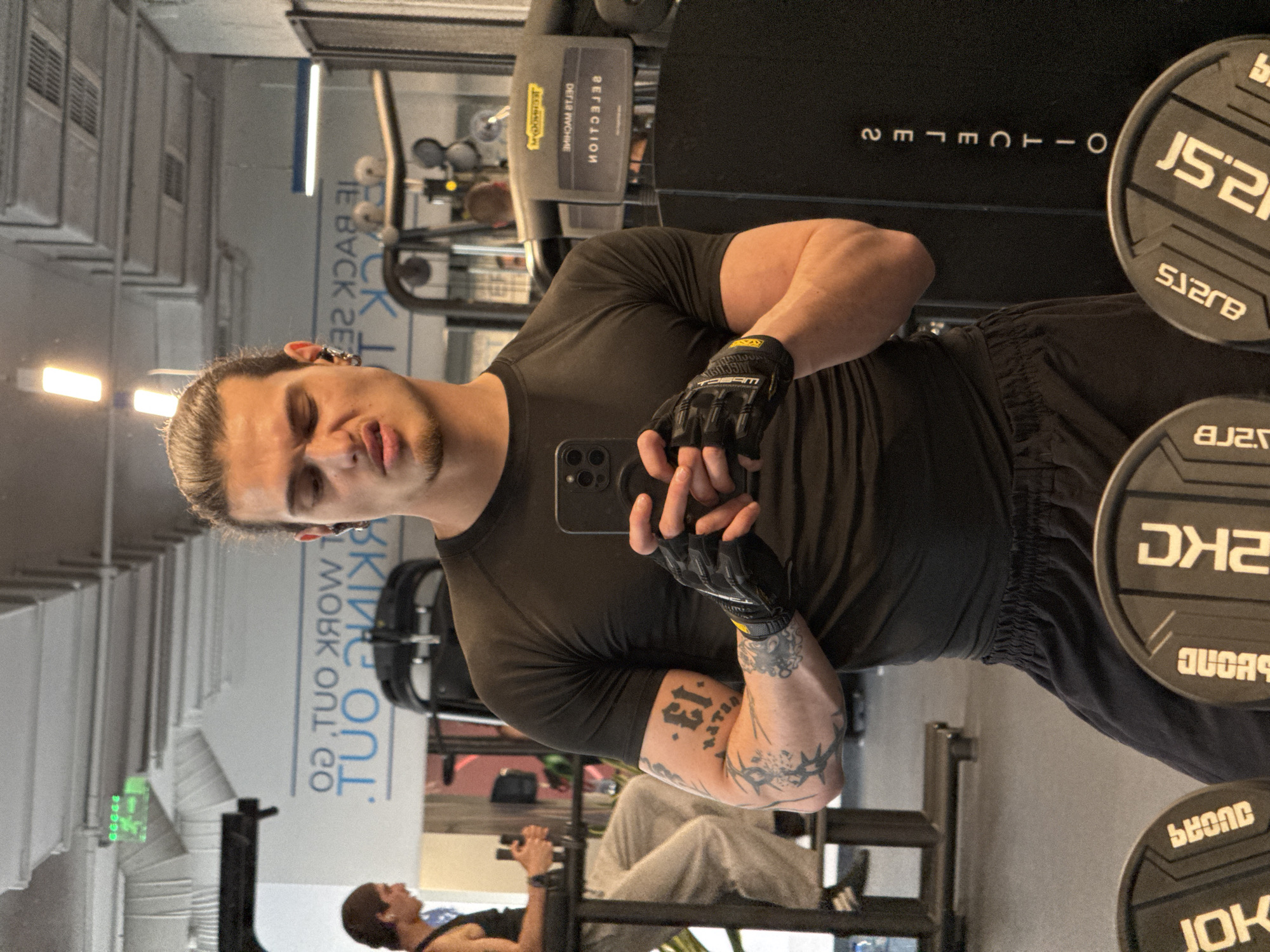 The image, rotated 90 degrees clockwise, captures a man taking a mirror selfie in an indoor gym in Sofia, Bulgaria. The man, with a focused expression and his hair tied back, wears a black short-sleeved t-shirt, dark athletic shorts or pants, and black fingerless workout gloves, possibly "MECHANIX WEAR" branded. His left arm displays prominent tattoos, including the number "13" and a detailed design resembling a thorny vine. He holds an iPhone with a triple-lens camera module, using it to capture his reflection.
The background, visible in the mirror, reveals various gym equipment. A large machine branded "TECHNOGYM SELECTION" (visible mirrored) stands behind him, along with several weight plates. One plate clearly shows "2.5KG," and another "2KG" (indicated by "2KC" in the OCR). Mirrored text on a wall or sign behind him reads "WORK OUT." Another person is partially visible in the reflection, also engaged in exercise, suggesting a typical gym environment during what appears to be daytime.