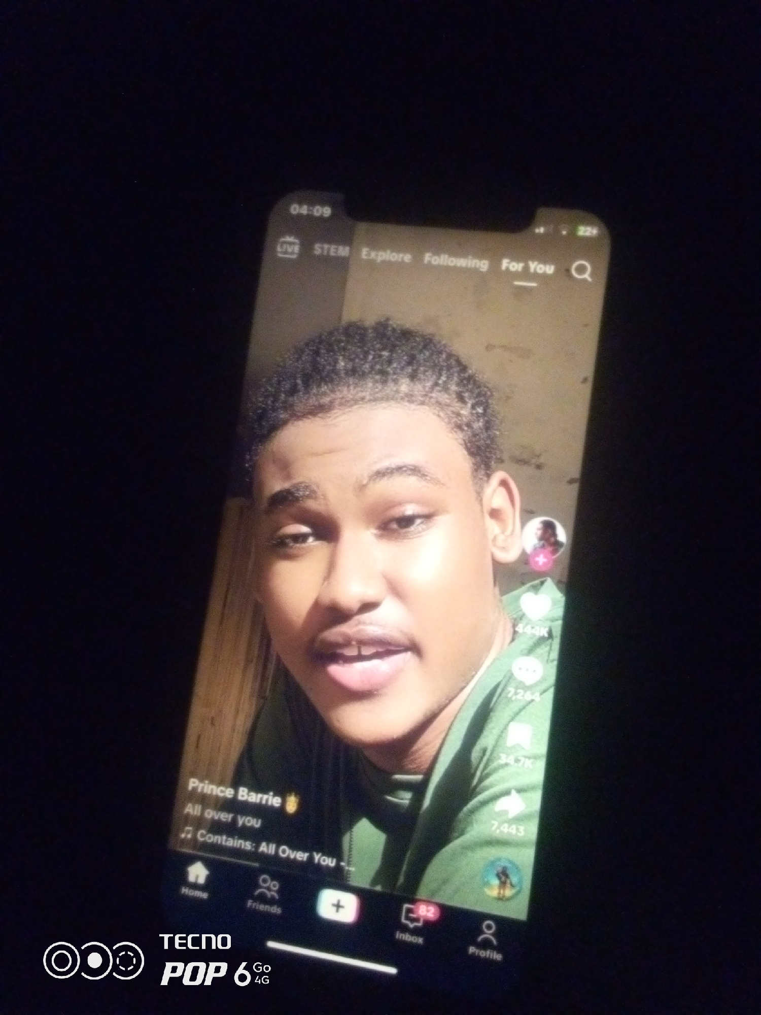 A close-up, overhead shot shows a smartphone screen displaying a social media video. The video features a young Black male with dark, curly hair and brown eyes, wearing a green t-shirt. His mouth is slightly open. The phone screen also shows various interface elements, including: a clock at 04:09, tabs for "LIVE," "STEM," "Explore," "Following," and "For You," a search icon, and on the right side, a profile picture of another person with a pink "+" sign and numbers indicating likes and shares (444K, 7,264, 34.7K, 7,443). At the bottom of the screen, there are navigation icons for "Home," "Friends," "Inbox" (with 82), and "Profile." The lower right corner of the image displays the "TECNO POP 6 4G" logo. The background is dark, suggesting the photo was taken in low light conditions, possibly indoors at night, in a location such as Howayi, Nigeria.