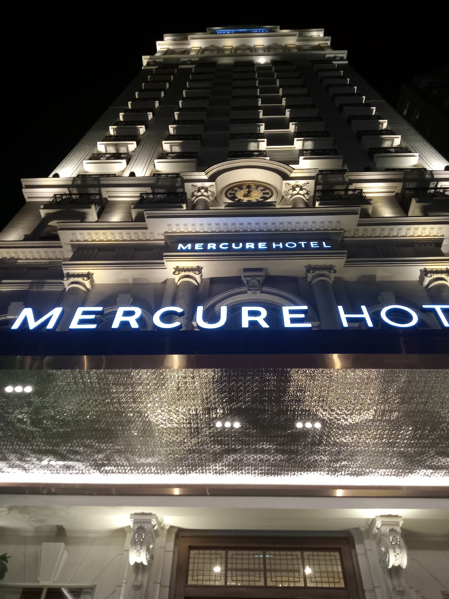 The image displays the illuminated exterior of the Mercure Hotel in Nha Trang, Vietnam, at night. The multi-story building rises against a dark, clear sky, with warm lighting highlighting its architectural details. The facade features numerous balconies with decorative railings, ornate columns, and cornices. A large, ornate clock face with golden Roman numerals is visible on an upper section of the building.
Two signs display the hotel's name: a smaller sign higher up reads "MERCURE HOTEL" in its entirety. Below it, a larger, brightly lit sign spells out "MERCURE HOTEL", though the final letters of "HOTEL" are partially cropped from the bottom of the frame. Below this sign is a textured, reflective surface, possibly a canopy or part of the building's entrance, adorned with embedded lights. The scene evokes a sense of grandeur and elegance, with the brightly lit building standing out in the nighttime setting.