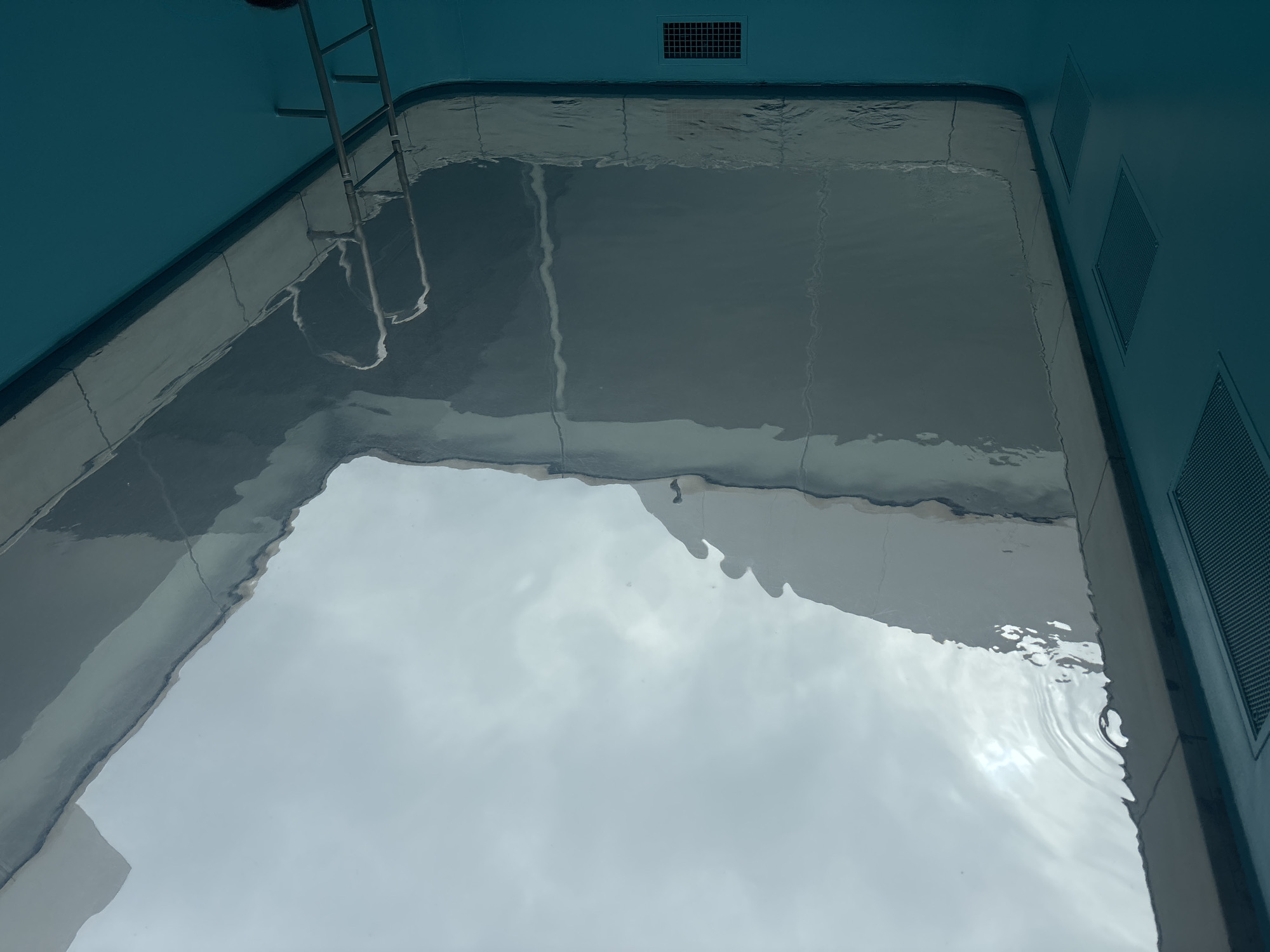 The image presents an overhead view looking down into a rectangular body of clear, tranquil water, enclosed by blue walls. This appears to be a pool or a large water feature within an indoor or covered setting, given the reflections.
In the top left corner, a metallic ladder with multiple rungs extends into the water, suggesting access for swimming or maintenance. Along the top blue wall, a single rectangular grated vent is visible. On the right-hand blue wall, three more identical grated vents are neatly aligned vertically.
The water's surface acts as a mirror, reflecting a bright, diffuse light source overhead. These reflections show broad, light-colored, cloud-like patterns, hinting at either a skylight illuminating an indoor space or an overcast sky if the area is open to the elements. Subtle ripples are visible on the water's surface, particularly towards the bottom right, suggesting slight movement or a recent disturbance. The floor of the pool, visible through the transparent water, is a dark gray and shows faint lines that might be tile seams or structural divisions.
No individuals are present in the scene, and no specific activity is taking place, contributing to a sense of calm and stillness. There is no visible text. The clean lines and functional structures suggest a modern facility, potentially a public bath, a hotel pool, or a feature in an architectural space, consistent with the location context of Kanazawa, Japan.