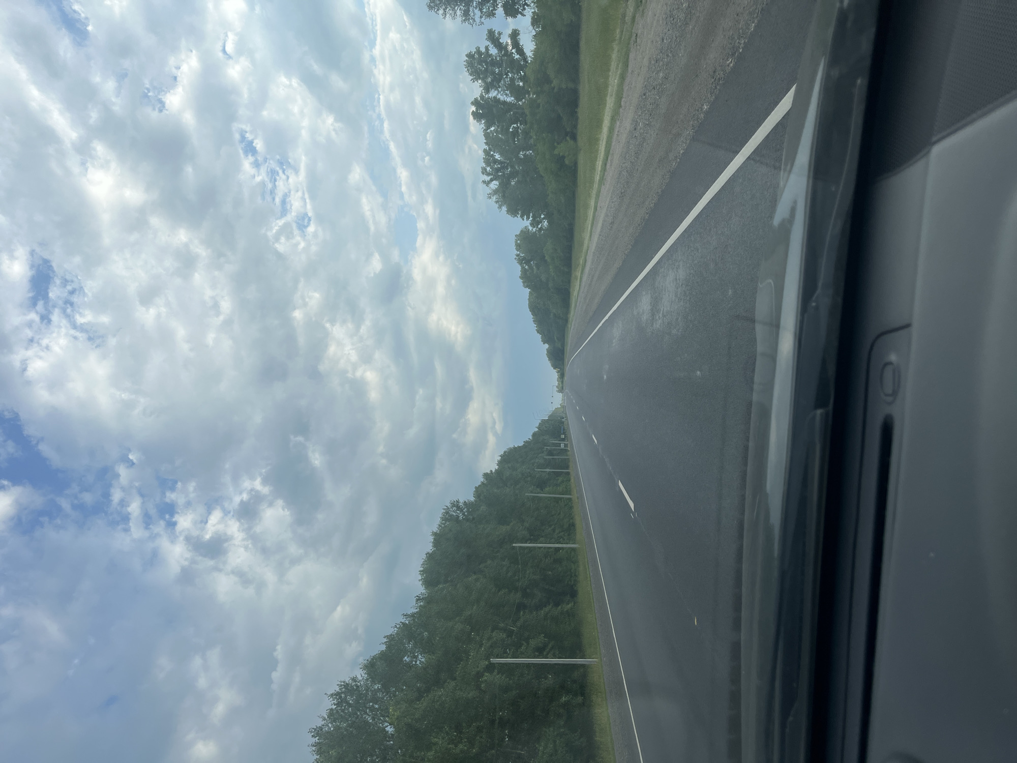 The media file depicts a view from inside a vehicle driving on a paved road. The road has a white line marking the lane. On the left side of the road, there is a dense line of green trees. The sky is filled with white and gray clouds, indicating overcast weather. On the right side of the frame, the interior of the vehicle is visible, showing dark gray panels and a white line that appears to be part of the car's interior design. The scene suggests a journey through a rural or suburban area. The time of day appears to be daytime, given the brightness of the sky. There is no visible text in the image.