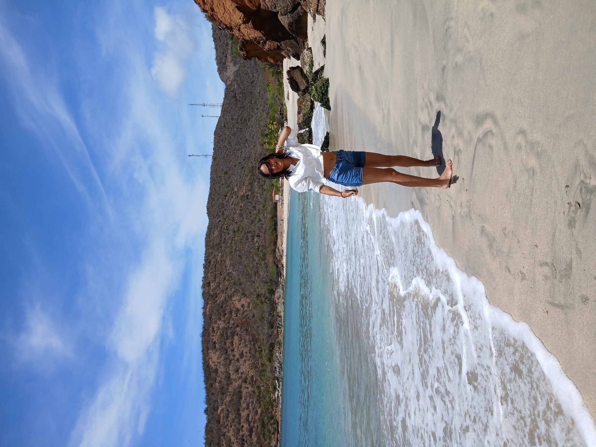 A vibrant, sunny daytime photograph captures a young woman posing on a sandy beach in Juan Griego, Venezuela. The woman stands towards the center-right of the frame, facing the camera with a slight smile. She is dressed in a white long-sleeved button-up shirt, tied at the waist to expose her midriff, and denim shorts. Sunglasses are pushed up onto her head, and she wears several bracelets on her wrists. Her bare feet are visible on the wet, light-colored sand, with a distinct shadow stretching to her left, indicating bright sunlight.
To the woman's left, the clear turquoise waters of the ocean gently meet the shore, creating white foamy waves that wash onto the beach. The sand is light tan and appears wet in areas where the waves have recently receded. Behind the woman, and extending across the horizon, are arid hills covered in sparse brown vegetation and rocky sections. On the crest of a distant hill, three distinct transmission towers are visible against the sky. The sky above is a bright, clear blue with a few scattered wispy white clouds. The scene portrays a serene coastal landscape under pleasant weather conditions. No visible text is present in the image.