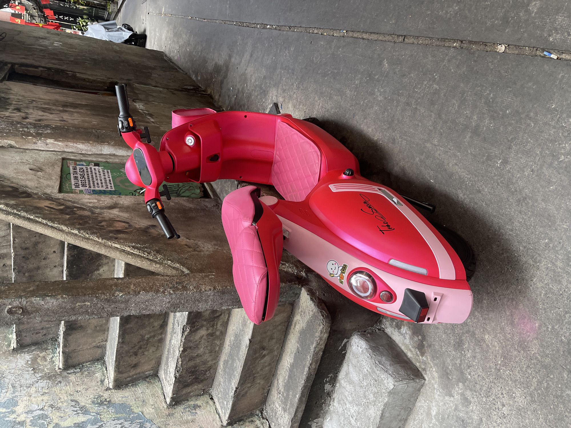 The image depicts a pink scooter parked on a concrete surface in what appears to be a narrow alleyway. The scooter has a shiny pink and light pink color scheme, a quilted seat, and decorative decals. It appears to be in Thủ Đức, Vietnam, as suggested by the Vietnamese text on a sign visible on the wall beside the scooter. The alleyway is narrow with weathered walls and concrete stairs.