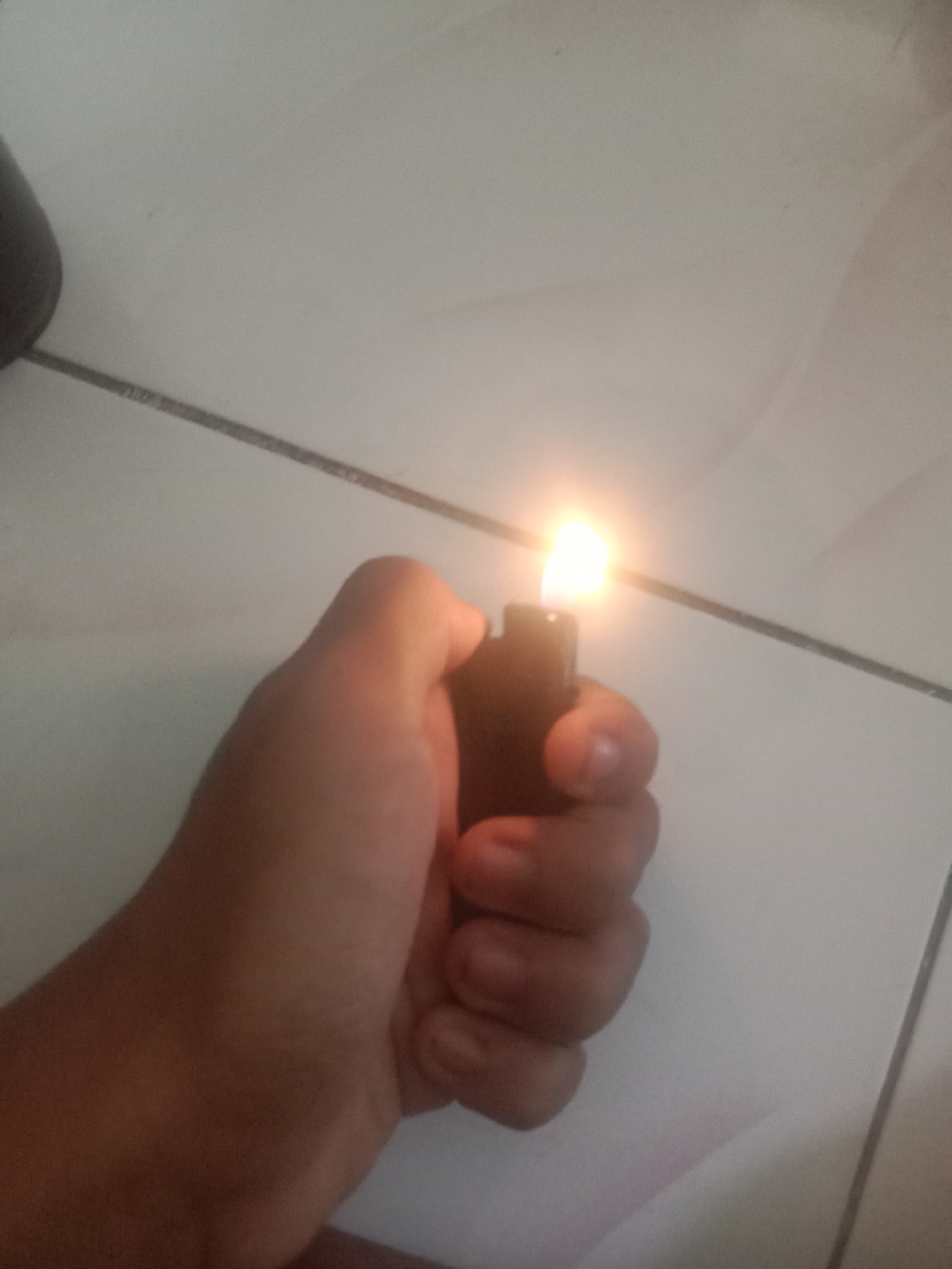 A hand is holding a lit lighter over a tiled floor. The lighter is black and the flame is a bright orange-yellow. The hand is light-skinned and appears to be that of an adult. The tiled floor is white with gray grout lines. The image is taken from a slightly elevated angle. There is no discernible information about the location in the city of Bumirejo, Indonesia.
