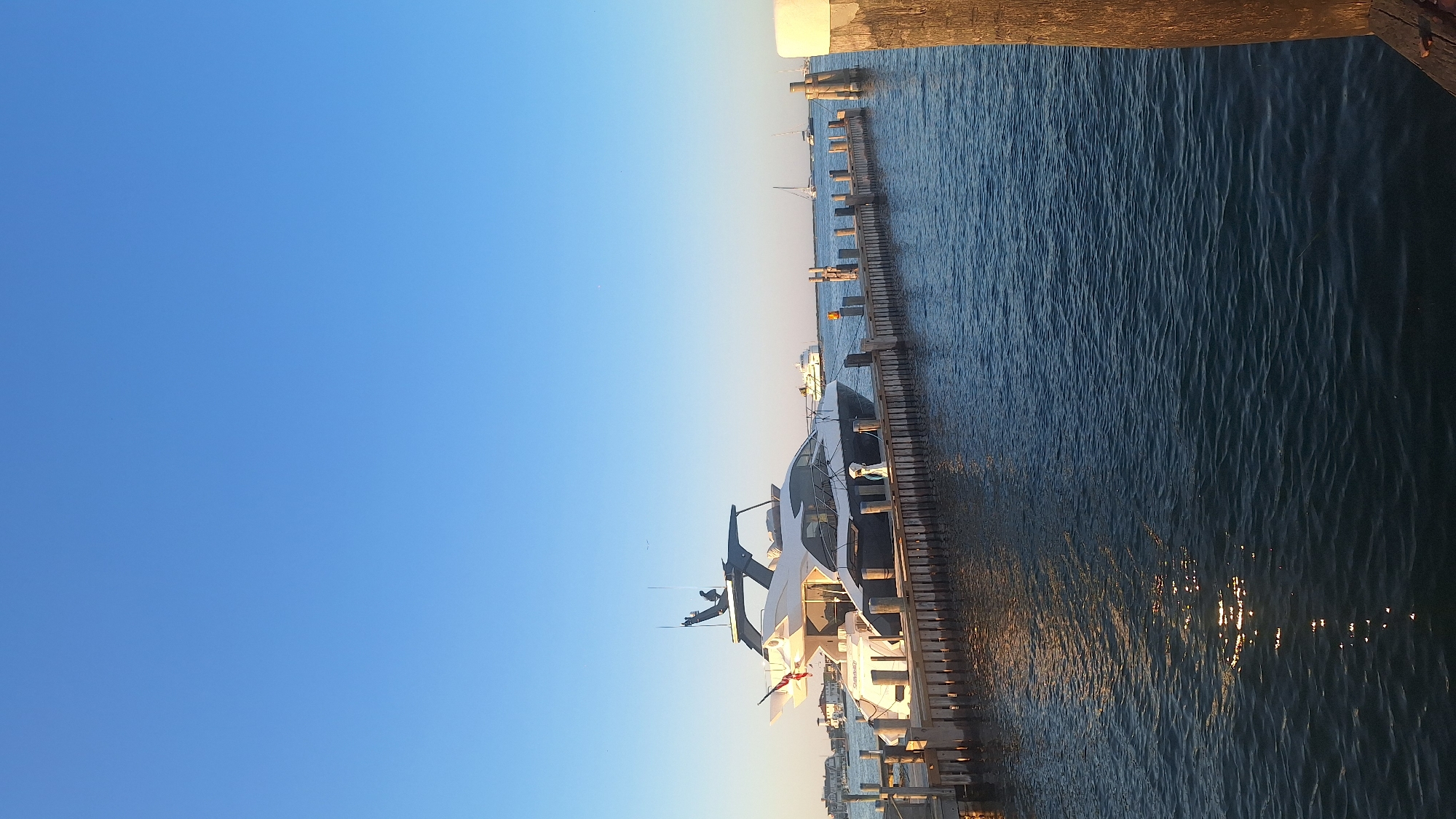 This image depicts a large, modern yacht docked at a wooden pier in Nantucket, Massachusetts. The yacht, predominantly white with dark accents, is the central focus of the scene. It is moored to a pier constructed of weathered wood pilings and planks. In the background, a clear blue sky dominates the upper portion of the frame, with a hint of the sun reflecting on the dark blue water. The water is calm, with gentle ripples and subtle reflections. Other vessels, including sailboats and a ferry, are visible in the distance, suggesting a busy harbor environment. Some buildings are visible on the shoreline in the far distance, hinting at the presence of a town or settlement. The overall scene conveys a sense of tranquility and luxury, characteristic of a waterfront location.