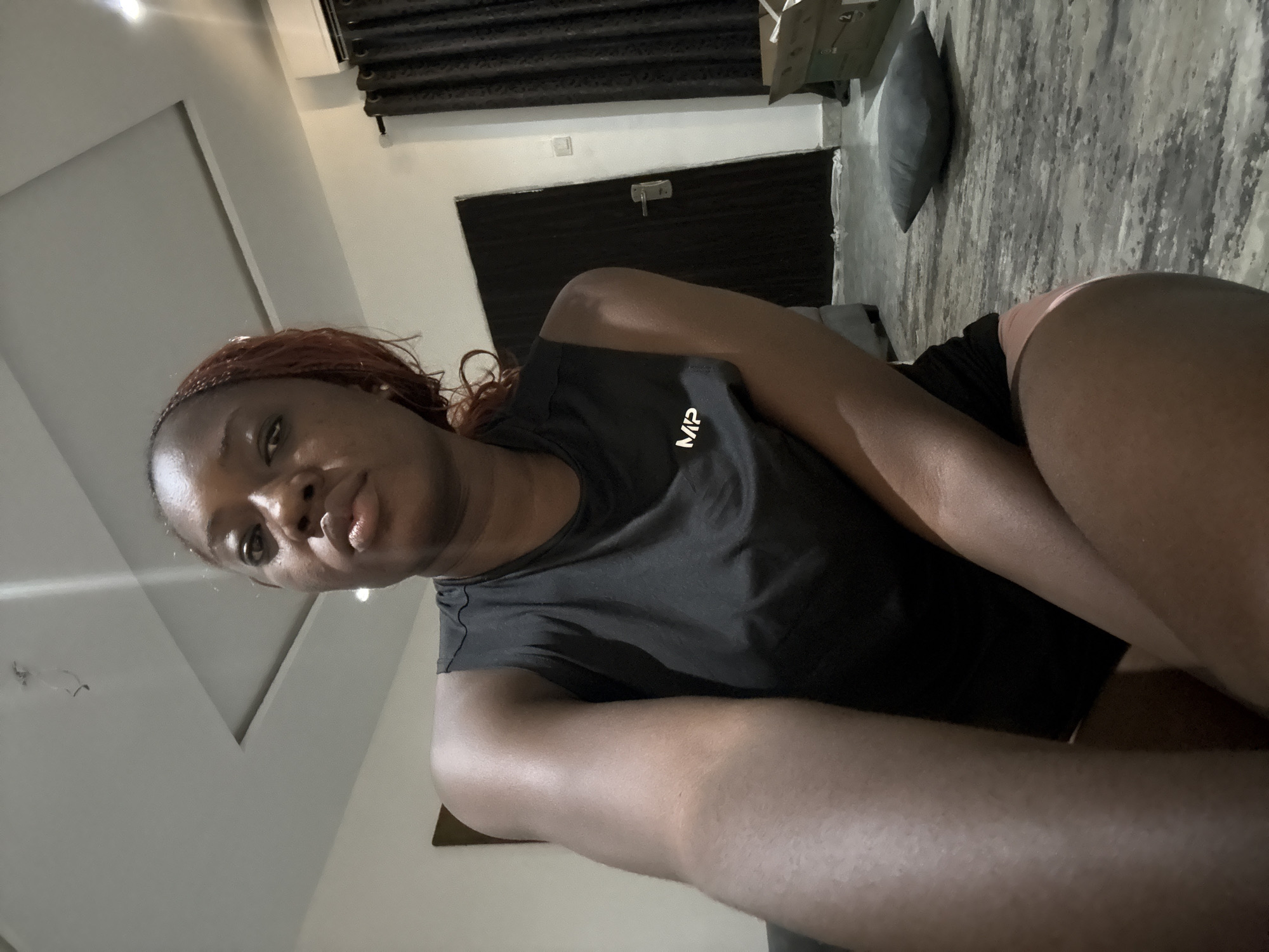 A woman with brown hair and dark skin is photographed from a low angle in an interior location. She is wearing a black tank top with "MP" printed on it, and dark shorts. The background includes a dark door with a silver latch, a patterned wall or floor covering, and a gray pillow. The ceiling has a modern design. The exact location is unclear, but presumed to be in Iddo Tudunwada, Nigeria, based on the provided information.