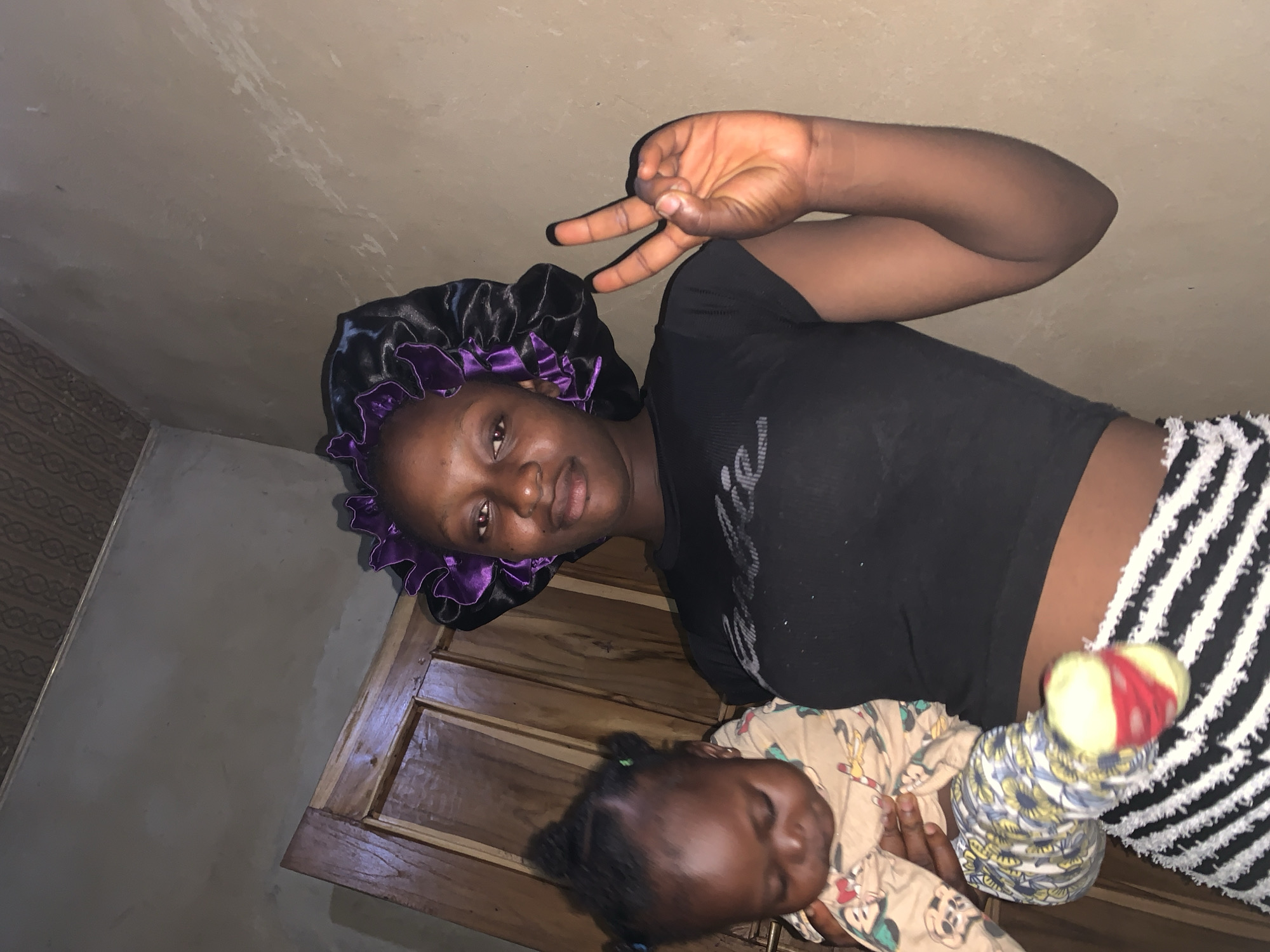 A young Black woman, wearing a black t-shirt, a black bonnet with purple trim, and striped pants, holds a baby wrapped in a light-colored outfit with cartoon characters and patterned leggings. The woman is looking at the camera and holding up two fingers in a peace sign with her right hand. The baby is facing away from the camera and appears to be looking down. They are standing in what appears to be a room with a wooden door in the background and a textured ceiling. The walls are a light brown color. This image was captured in Agban, Nigeria.