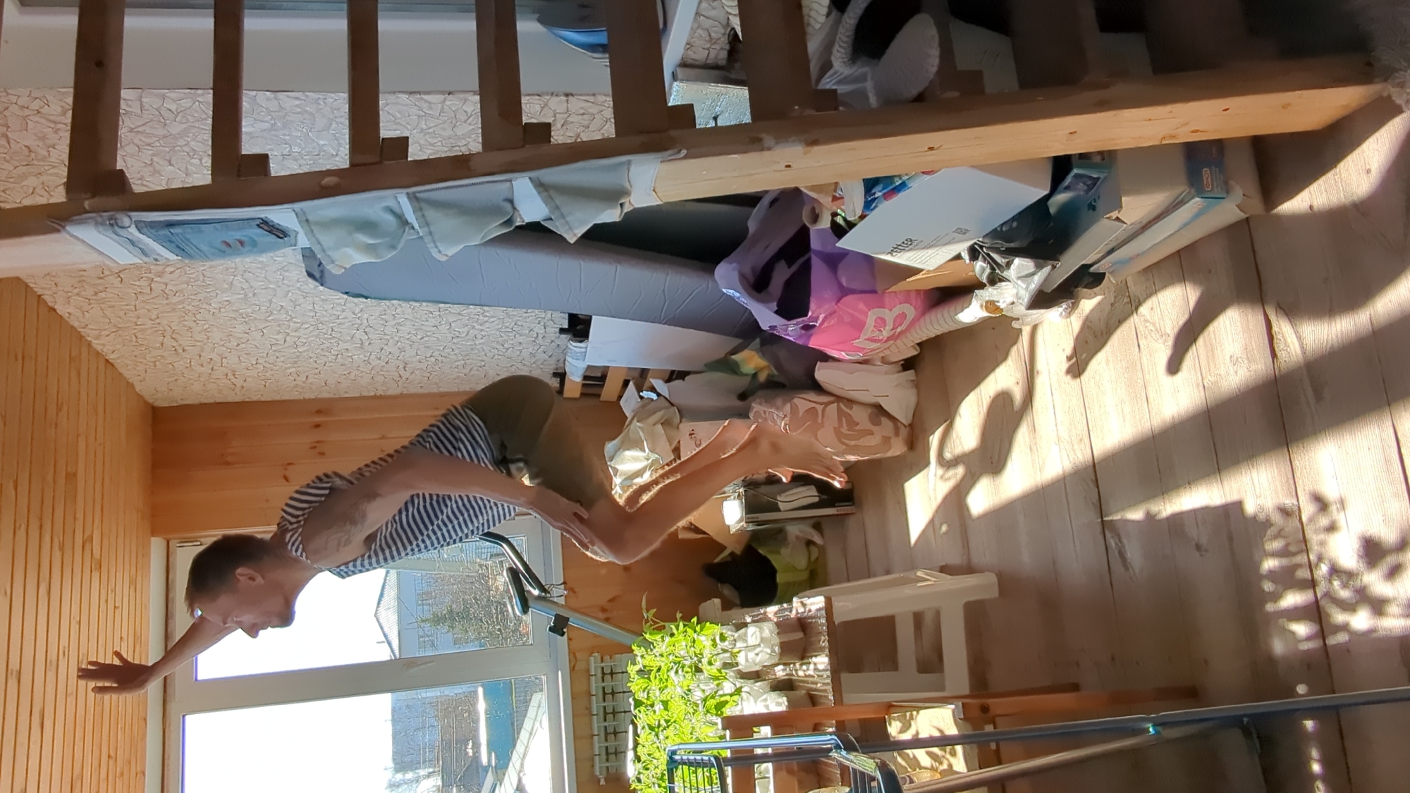 The image portrays an indoor residential scene, likely a room or attic space in Ulyanovsk, Russia, illuminated by strong natural light. A man is captured in a balanced pose, performing an exercise or yoga movement. He wears a striped short-sleeved shirt and olive green shorts, and is barefoot, with a focused expression as he holds his position with outstretched arms.
The room features a wooden paneled wall to the left and wooden floorboards on the right, where sharp shadows from objects and sunlight create distinct patterns, indicating it is daytime with clear weather. The ceiling is covered with patterned wallpaper. A large window behind the man provides a glimpse of outdoor buildings and trees.
In the background, a wooden staircase railing spans across the upper portion of the frame. Beneath it, a gray mattress or sofa bed is surrounded by various cluttered items, including boxes, bags, and blankets. Text "ZETTER" is visible on one box, and a large "B" is seen on a pink bag. An exercise bike and a shelf with potted plants are also present in the mid-ground, contributing to the lived-in and somewhat busy atmosphere of the room.