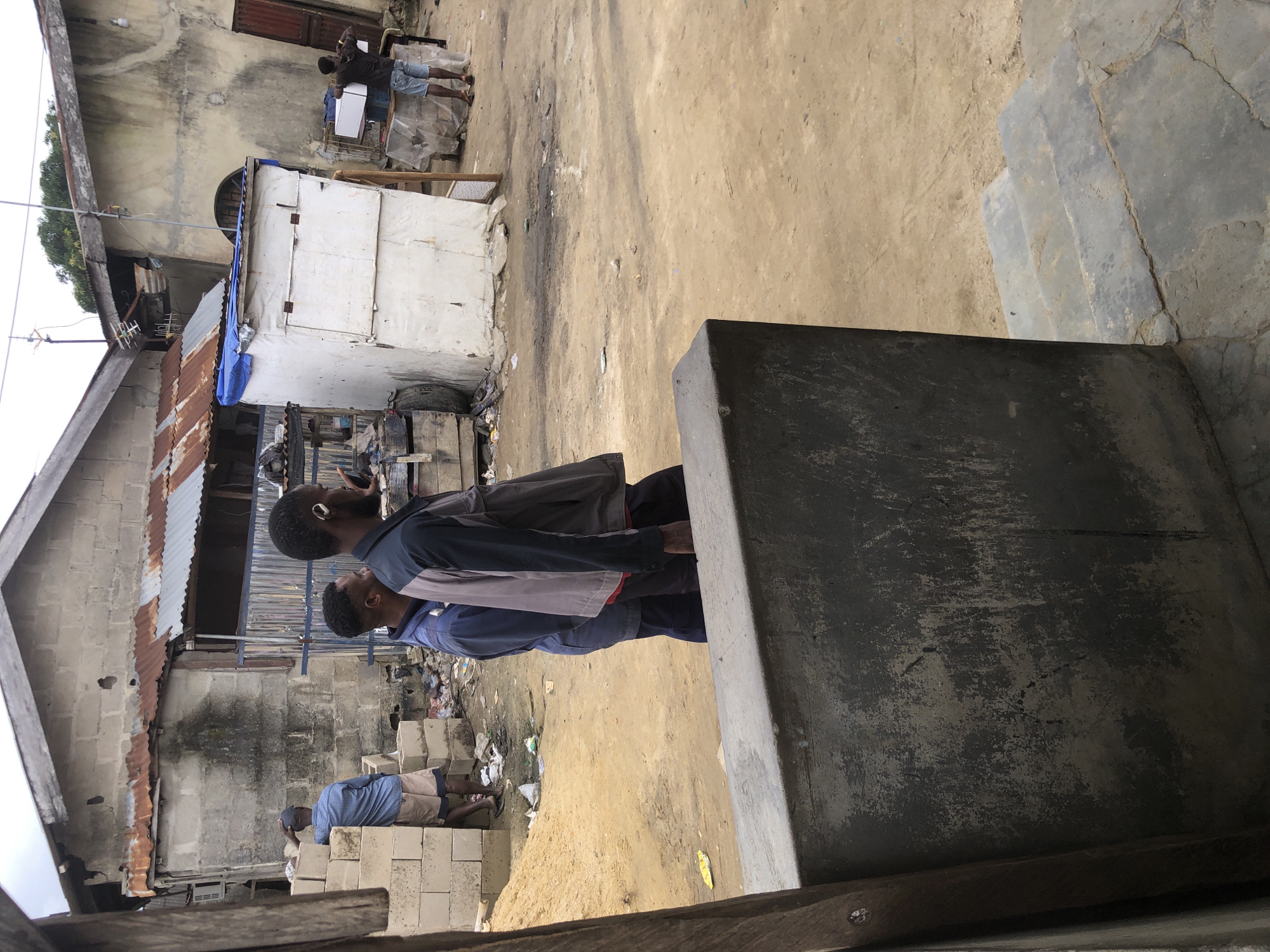 A street scene in Ado, Nigeria, depicts two men in the foreground, walking from left to right. The man on the left wears a blue jacket and dark pants, while the man on the right wears a gray and dark-colored jacket with dark pants and a white earpiece. Behind them, another man in a blue shirt and shorts is seen near a stack of concrete blocks. The background features a cluster of buildings with corrugated metal roofs and weathered walls, suggesting a modest urban or semi-urban environment. The ground is a mix of dirt and sand, with some scattered debris. To the right, a small white structure with a blue tarp roof stands near another building. A man is visible in the distance carrying a white box. The overall impression is a snapshot of daily life in a Nigerian city, characterized by its architecture, the attire of the people, and the state of the environment.