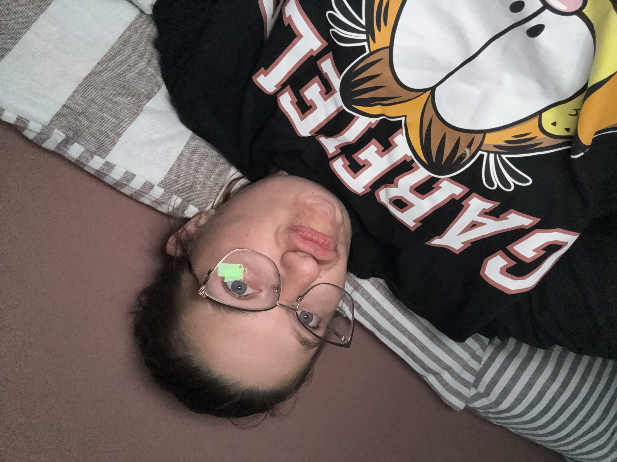 A woman with glasses is lying down on a bed. She is wearing a black shirt with a cartoon graphic and the word "GARFIELD" in white and maroon lettering. The bed has a striped pillowcase. The location of the image is not specified, so it is not possible to determine if this photo was taken in Sofia, Bulgaria, based on the image alone.