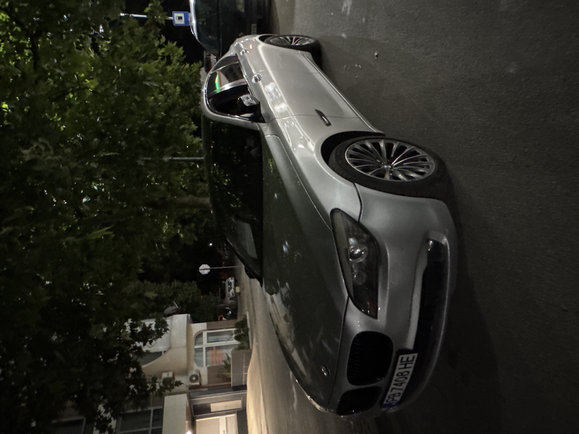 A silver BMW 7 Series sedan is parked on a street at night in Plovdiv, Bulgaria. The car's license plate reads "PB 7408 HE". A dark-colored car is parked behind it. The scene is lit by streetlights and the headlights of the cars. Trees with dark green leaves overhang the street, and a building is visible on the left side of the image. The asphalt road is wet, indicating recent rain.