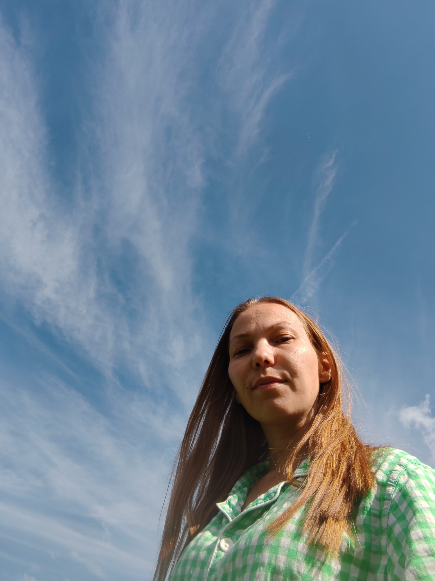 A low-angle, close-up selfie shows a woman with long, reddish-brown hair against a backdrop of a bright blue sky with wispy white clouds. She is wearing a green and white checkered shirt. The woman's face is turned slightly upwards, and she has a neutral expression. The image appears to be taken outdoors, possibly in Чухлэм, Russia, given the clear sky and natural light.