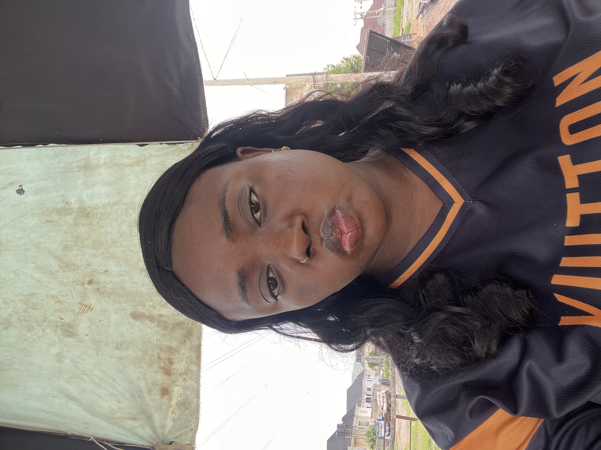 A medium shot of a Black woman with dark, wavy hair wearing a dark blue jersey with orange lettering ("VUITTON") and trim. She has a nose ring and appears to be puckering her lips. The photo seems to be taken outdoors under an awning with a tent-like fabric. In the background, there are utility poles, buildings, and vegetation, suggesting a residential or commercial area. There is no specific information to confirm the location within Abuja, Nigeria.