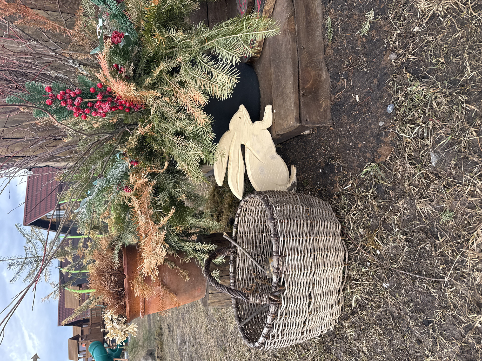 The scene depicts an outdoor decorative arrangement in Tyulyuk, Russia. In the foreground, a large, light-colored wicker basket with a dark, twisted handle rests on a patch of dry, brown grass mixed with bare soil. The basket contains several small, dry twigs. To its immediate right, a pale wooden cut-out of a rabbit, facing left, is positioned partially on the ground and partially on a dark brown, multi-tiered wooden platform. On this platform, a black pot holds a dense arrangement of green fir branches, some exhibiting a yellowish-brown tint, along with a prominent cluster of red berries, possibly artificial, coated in a light, frosty substance. Behind this, to the left, a terracotta-colored pot contains dried, reddish-brown foliage. Numerous thin, dark, bare branches extend upwards from behind these potted arrangements. The background reveals parts of wooden structures, including what appears to be a fence or wall, and several houses with dark red gabled roofs and lighter-colored walls. To the far left background, a children's playground structure with a green slide is visible. The sky overhead is overcast. No individuals are present in the image.