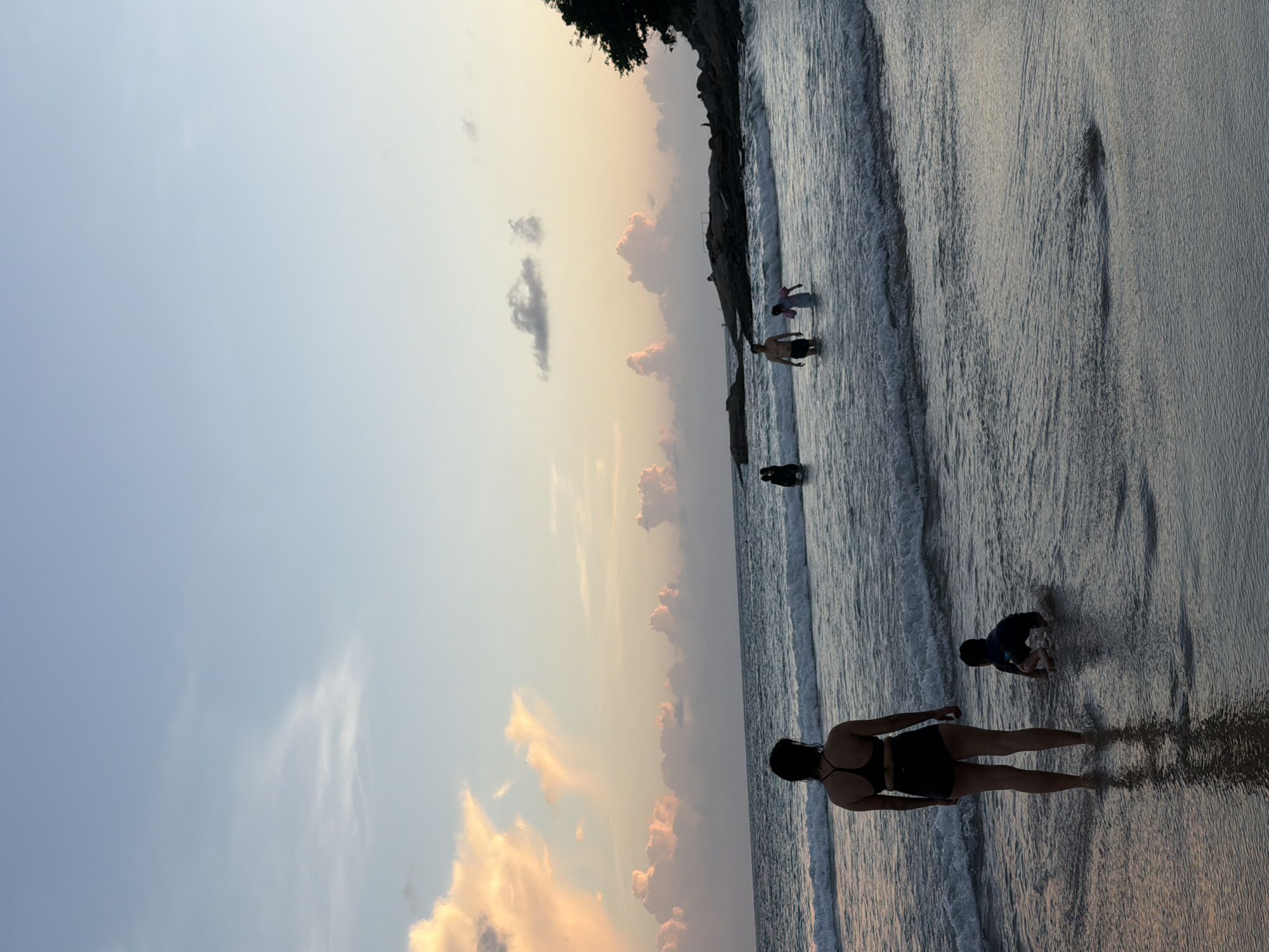 The image depicts a serene beach scene at sunset in Rawai, Thailand. The sky is painted with soft hues of orange and pink, indicating the late afternoon or early evening. Gentle waves lap at the shore, creating a calming rhythm.
Several people are present on the beach, enjoying the tranquil atmosphere. In the foreground, a woman stands facing the water, her silhouette outlined against the setting sun. Further down the shore, a child is seen playing in the shallow water, crouching and interacting with the waves. Other figures are scattered along the waterline, some wading and others walking.
The composition highlights the vastness of the ocean and the sky, with the shoreline acting as a natural division. The presence of a few scattered clouds adds texture to the sky, while the overall lighting suggests a peaceful and relaxing environment. The dark silhouette of a tree is visible on a rocky outcrop in the distance, adding a touch of natural beauty to the landscape. The image evokes a sense of calm and enjoyment of a beautiful day at the beach. There is no visible text in the image.