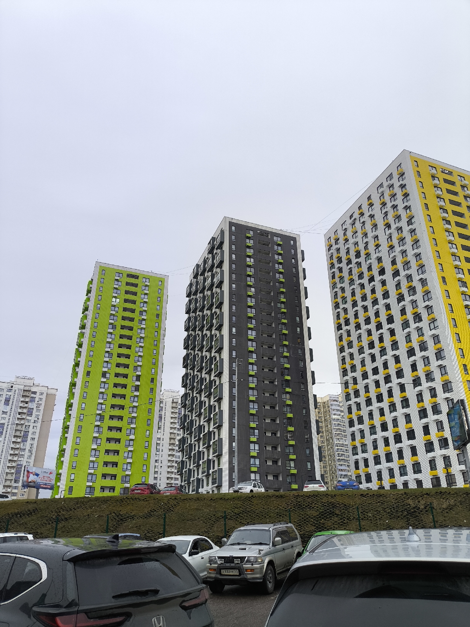 The image depicts a street scene in an urban area. Three brightly colored high-rise buildings, painted in green, black, and yellow respectively, dominate the background. A green embankment separates the buildings from a row of parked cars in the foreground. The sky is overcast, suggesting a cloudy day. The license plate on one of the parked cars reads "Y733 PE 99".