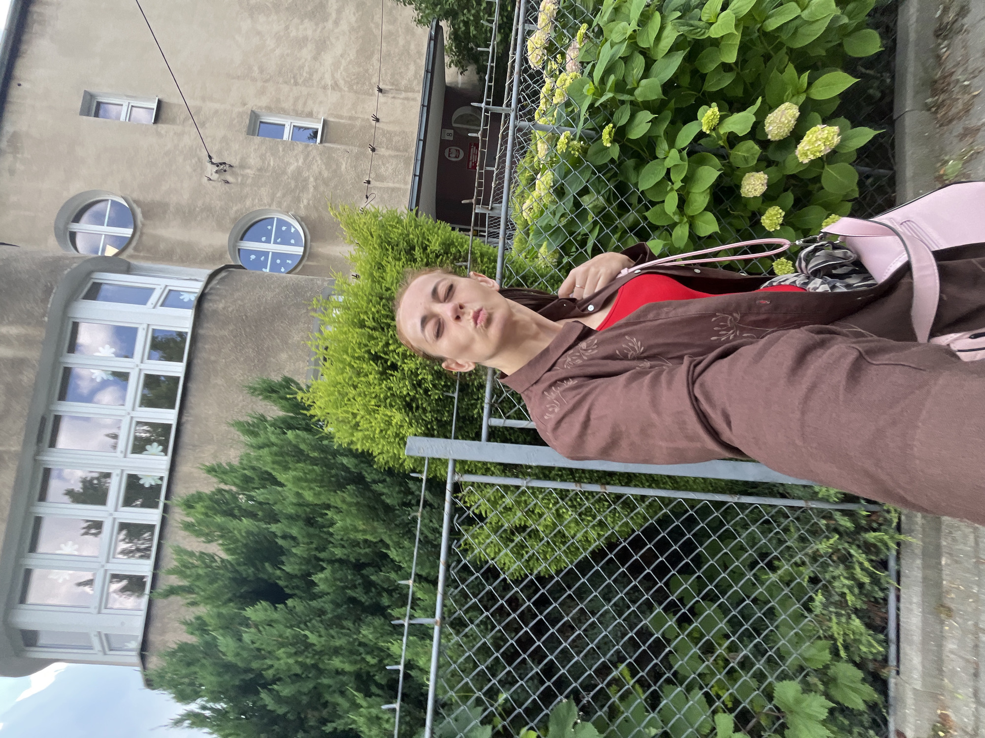 In this portrait-oriented image, a young woman with blonde hair pulled back is facing the camera, making a kissing face. She is wearing a red top and a brown jacket with a floral pattern. A pink purse is slung over her shoulder. She is standing outdoors in front of a chain-link fence, with lush green foliage and flowering bushes behind it. To her left, part of a building is visible. The building has a textured, light-colored exterior and features several windows, including some circular ones with crossbars. The image appears to have been taken during the daytime, under overcast skies, as suggested by the soft lighting and muted colors. The overall mood is casual and playful, with the woman's expression conveying a sense of lightheartedness. There is no discernible text within the image.