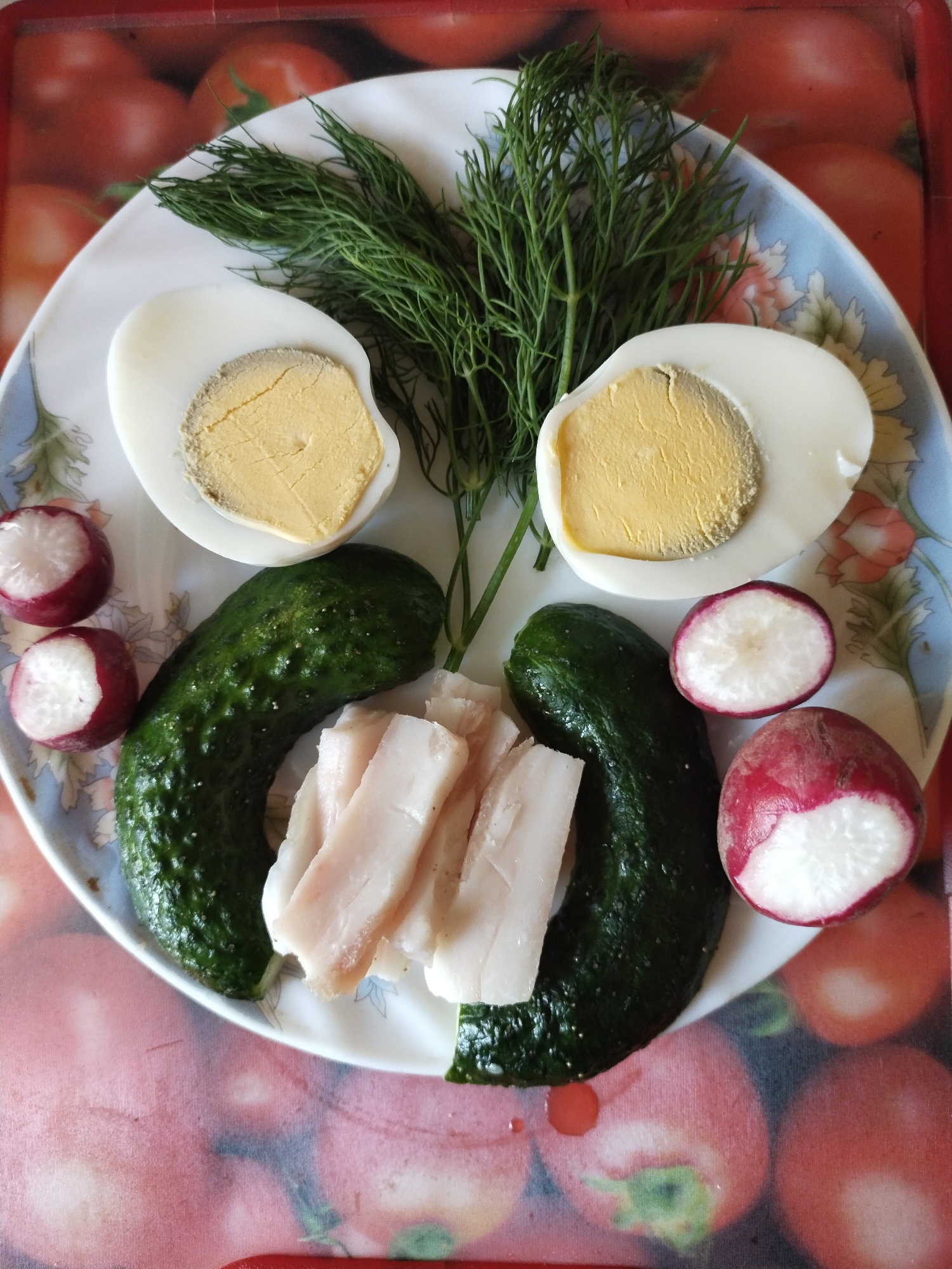 A plate of food, including two halves of a hard-boiled egg, a bunch of dill, two cucumbers, several pieces of white fish, and four radishes, is placed on a red tray with a pattern of red tomatoes. The scene is indoors.